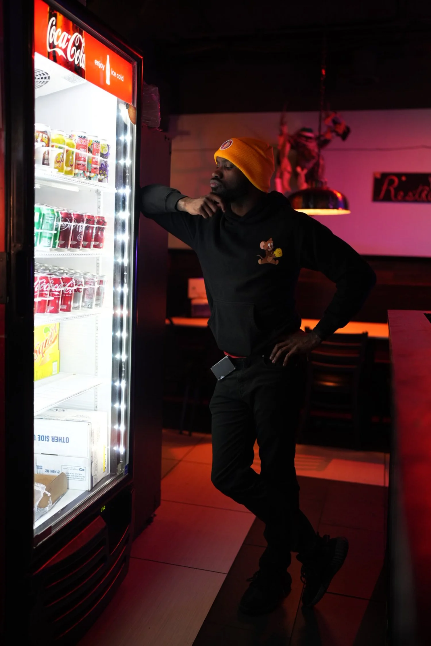 A man wearing a yellow beanie, black hoodie with a cartoon bear logo, and black pants is standing next to a Coca-Cola vending machine in a dimly lit restaurant or bar, with pink and red lighting in the background.