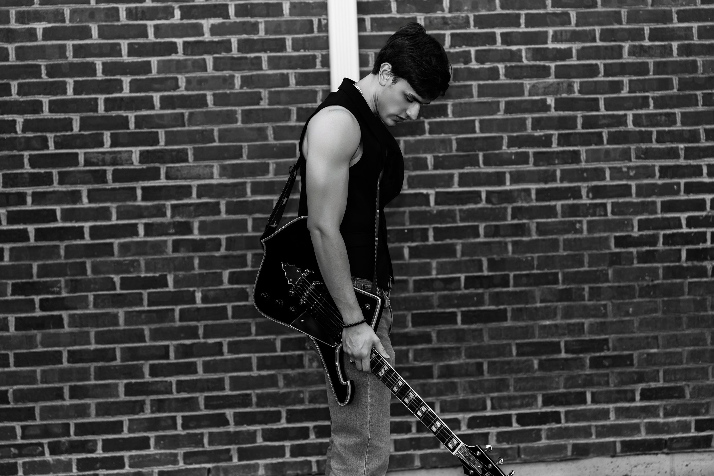 A young man with dark hair, wearing a sleeveless black vest and jeans, stands against a brick wall, holding an electric guitar with the headstock touching the ground and looking down.