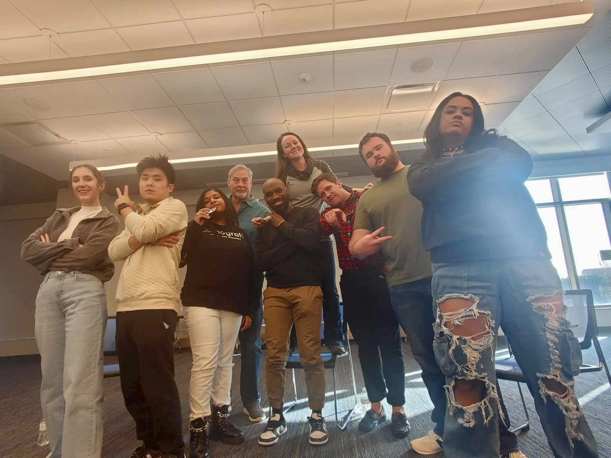 A diverse group of nine young adults posing together in a room with chairs and large windows, some making peace signs or crossing arms, all smiling or looking confident.