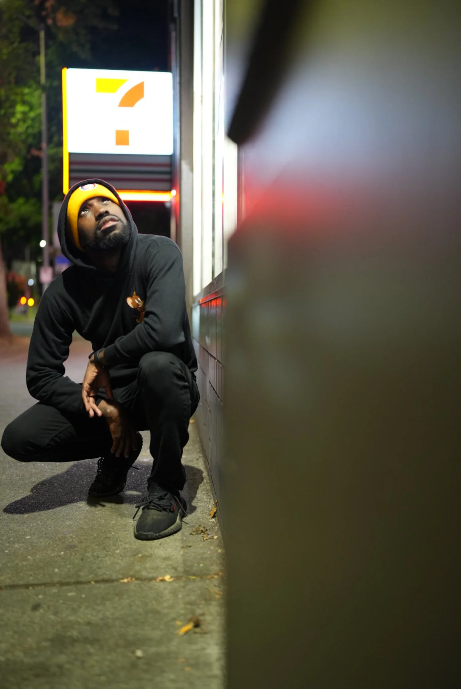 A man crouching on a sidewalk in front of a wall, looking up, wearing a black hoodie with a logo, black pants, black shoes, and a yellow beanie, near a brightly lit sign and a metal barrier.