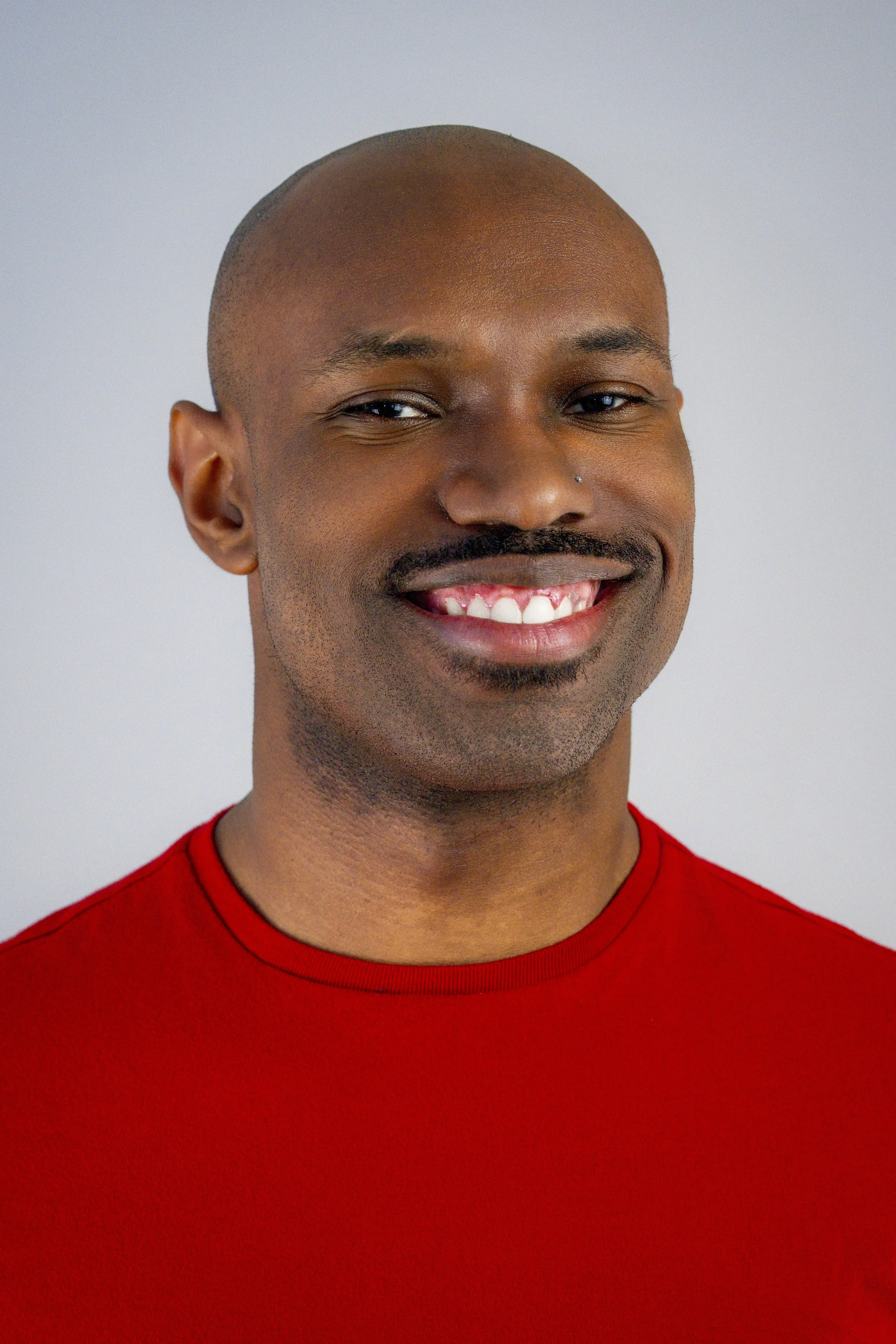 Close-up portrait of a smiling Black man with a shaved head, mustache, and beard, wearing a red shirt against a plain light background.