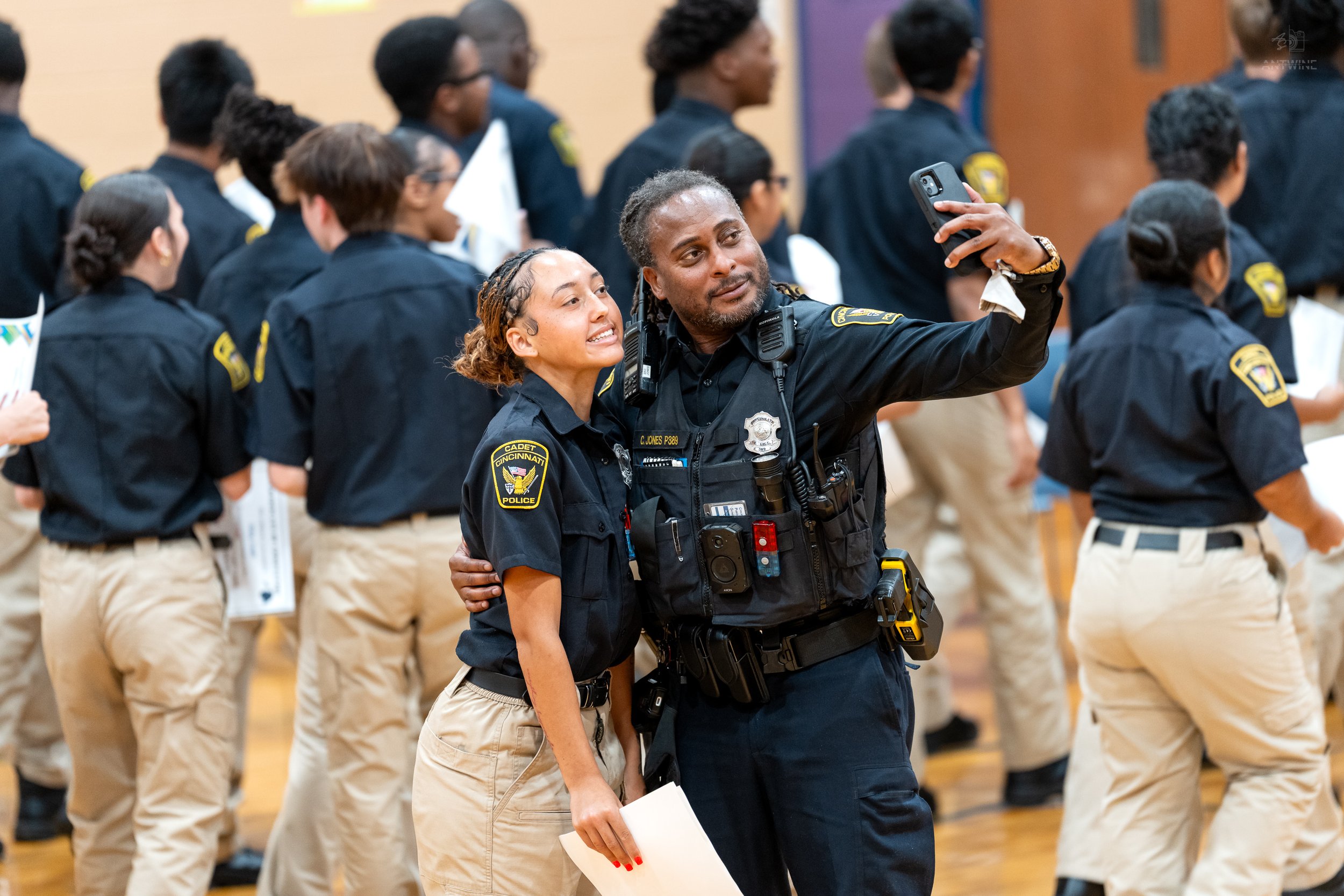 Two police officers, a man and a woman, taking a selfie together at a gathering of officers in uniform, with several other officers standing in formation behind them.