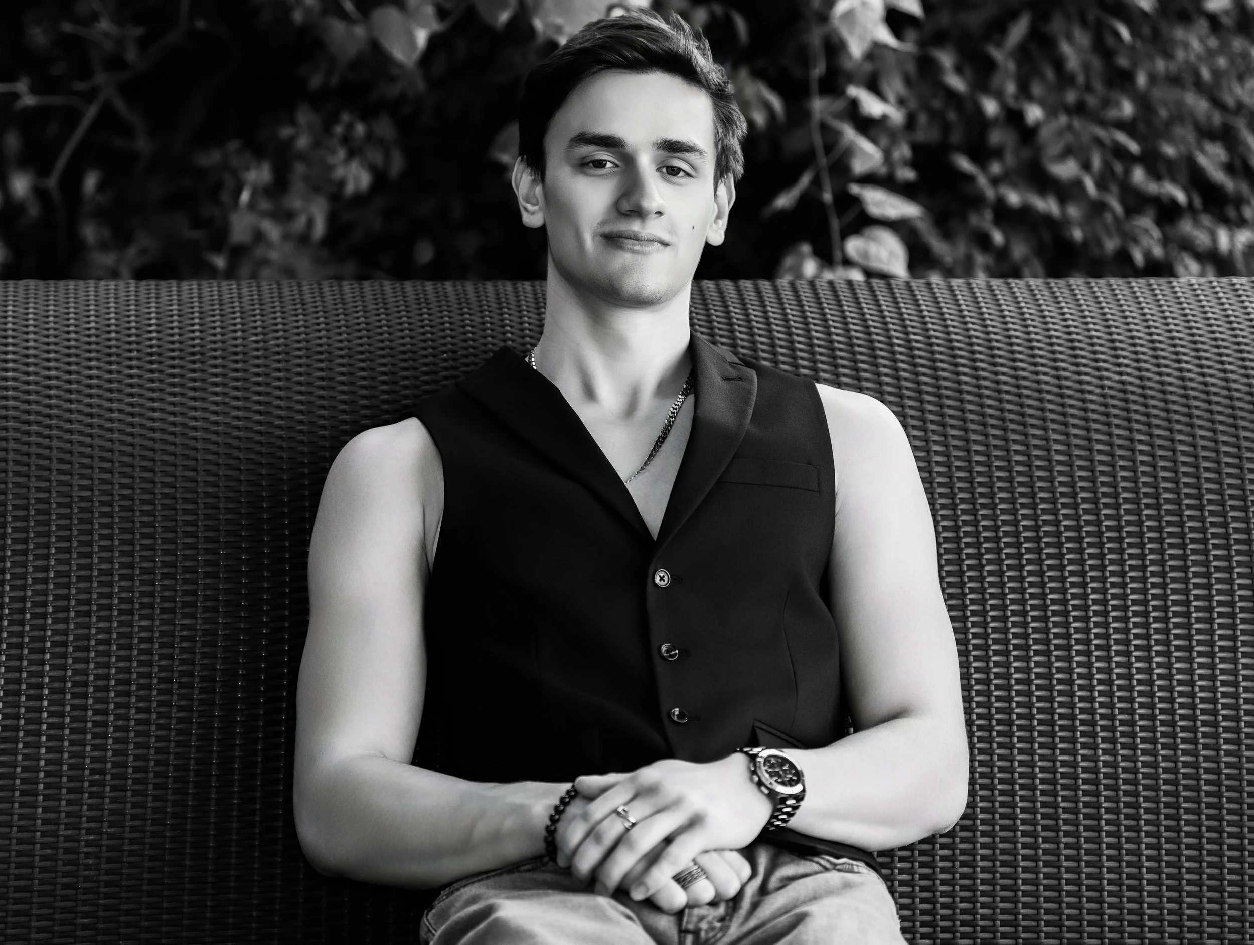 A young man with dark hair sitting on a wicker bench outdoors. He is wearing a sleeveless black shirt, watch, and accessories. The background consists of foliage.