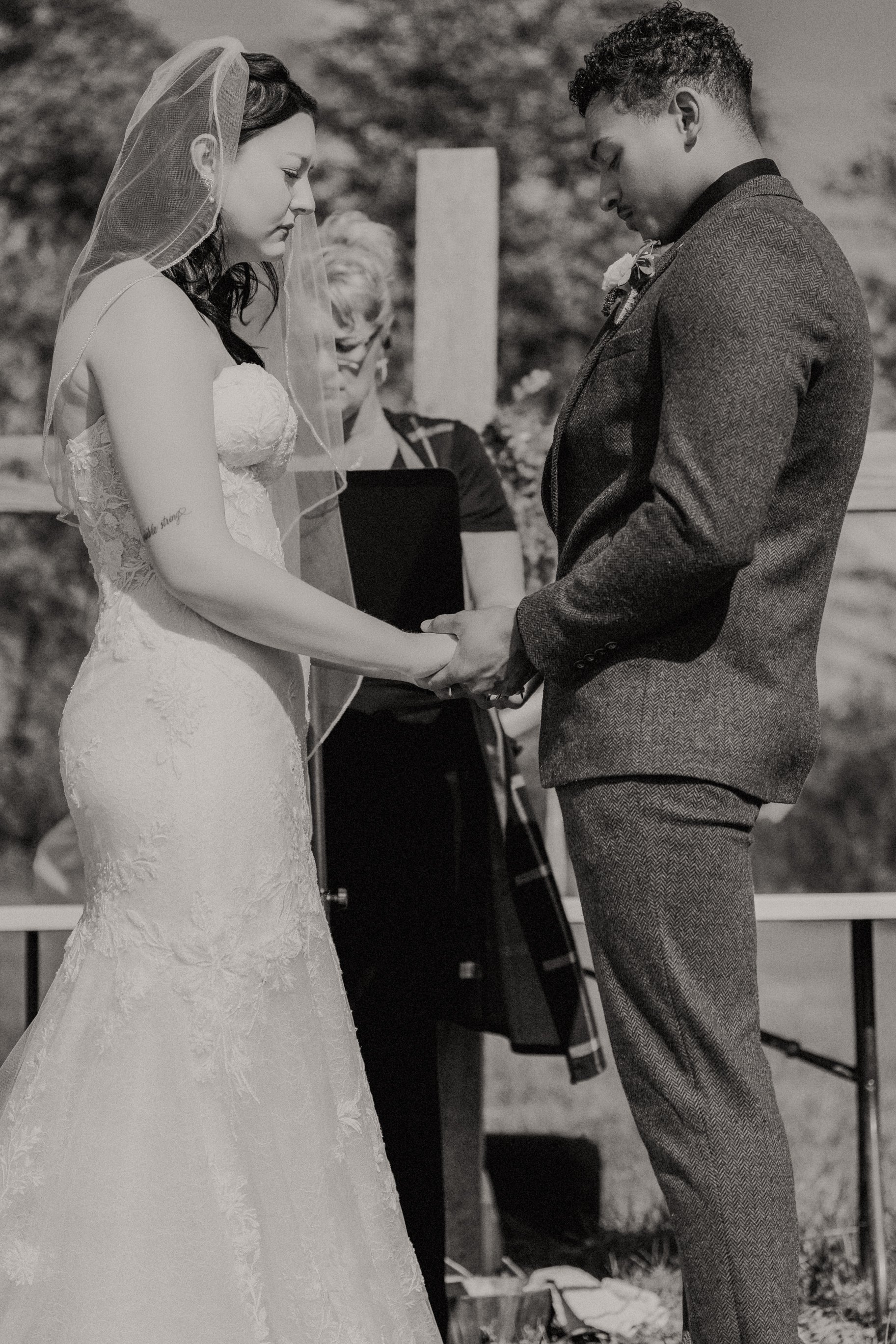 A black and white photograph of a wedding ceremony, with a bride and groom holding hands and bowing their heads, standing outdoors.