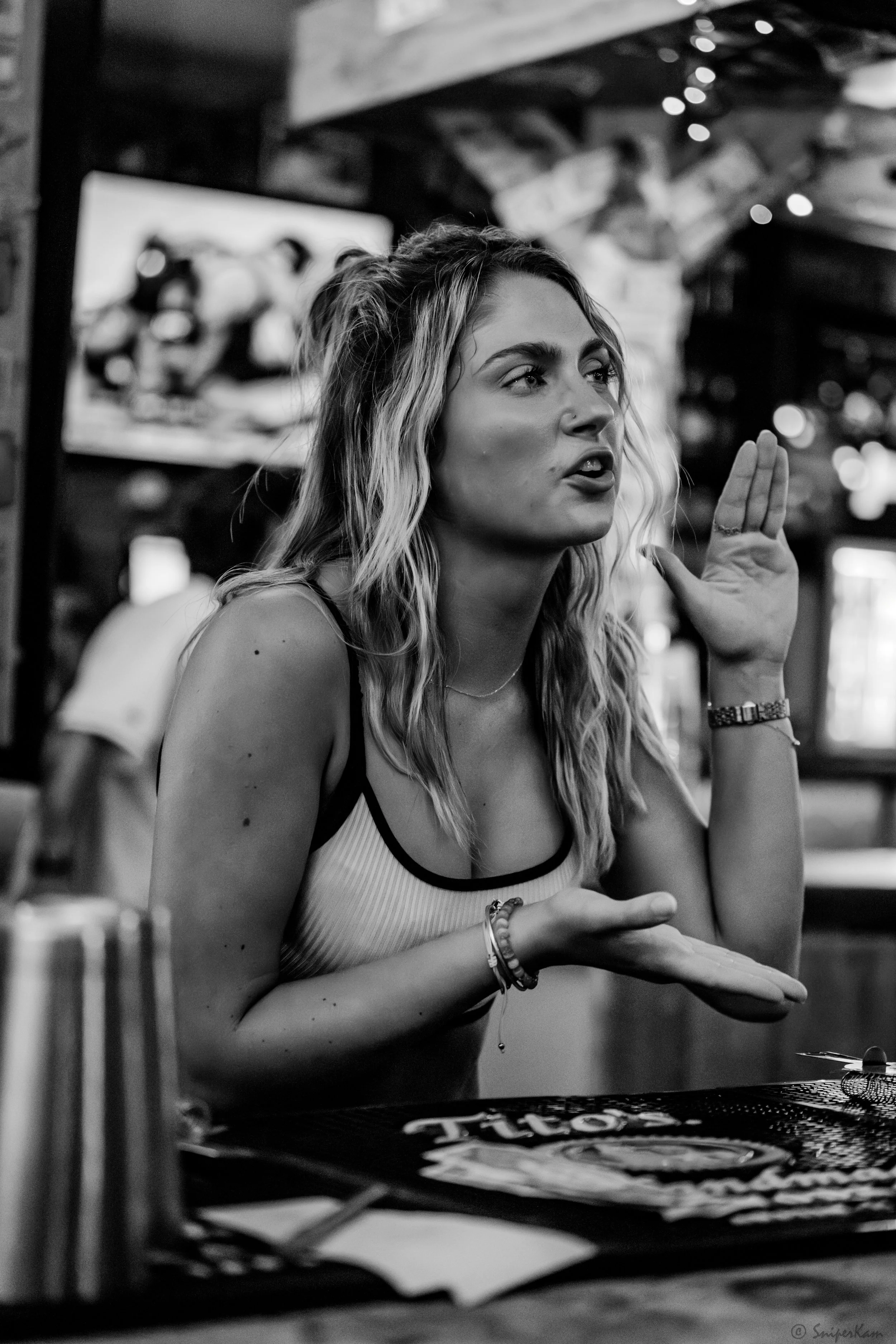 A woman with wavy hair and a sleeveless top engaging in conversation at a restaurant or bar, gesturing with her right hand, with a background displaying a television and string lights.