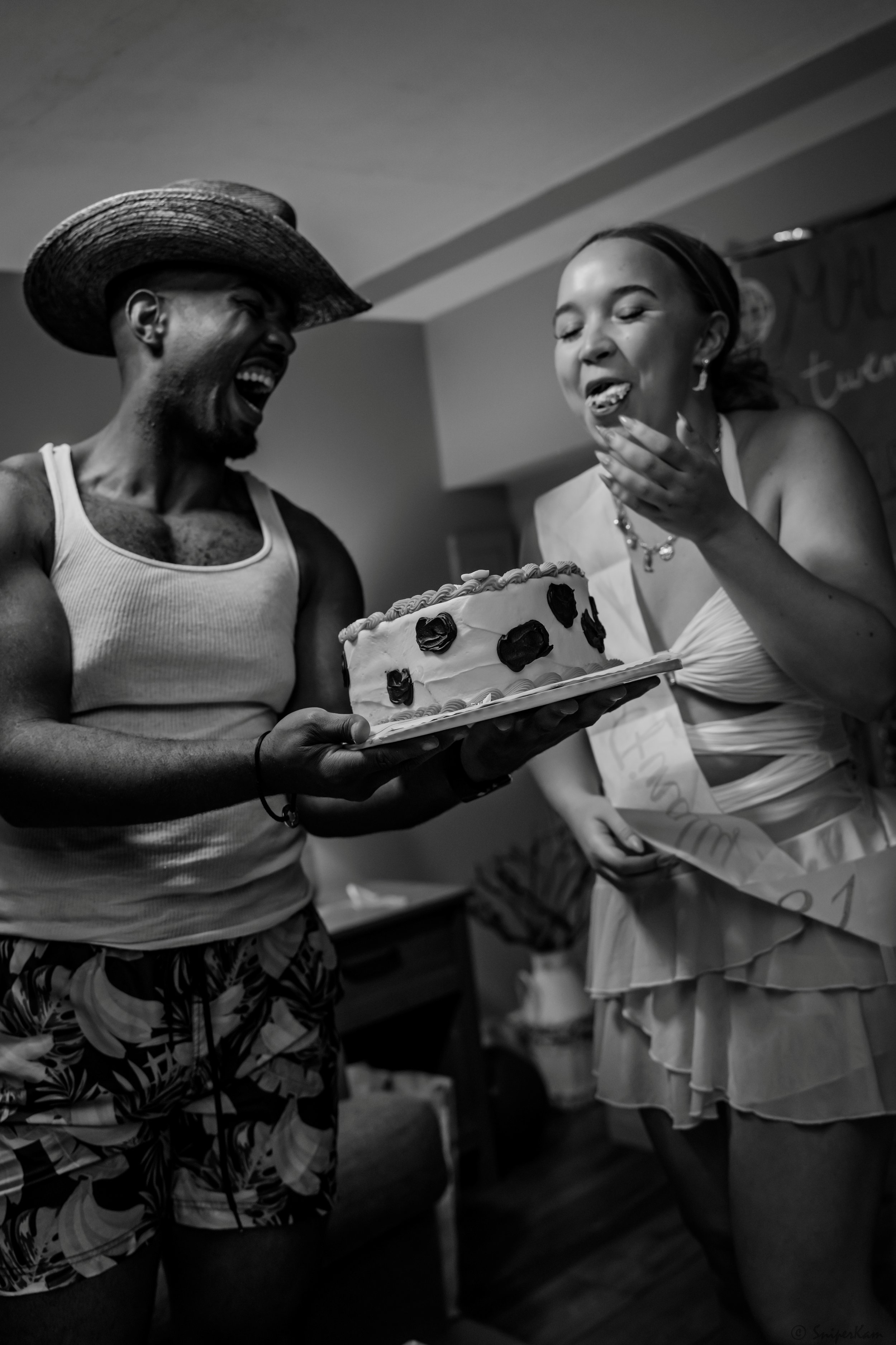 A man in a tank top and straw hat presenting a cake to a woman with a sash, both smiling and laughing at a celebration.