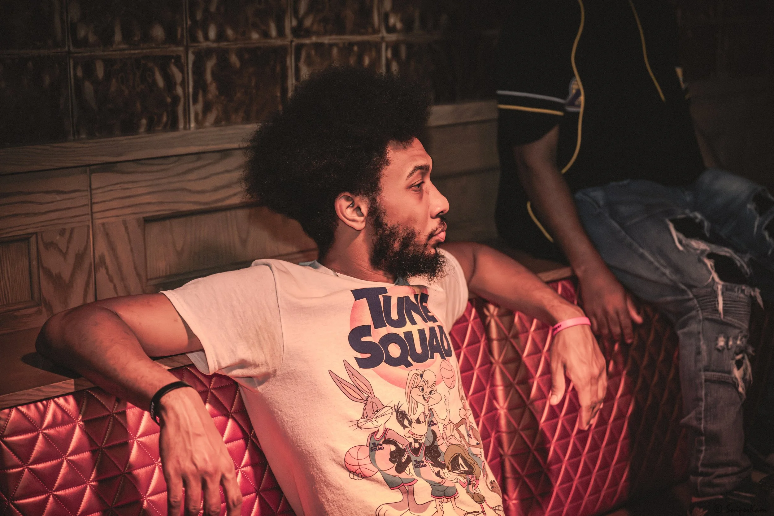 A young man with an afro hairstyle and beard is sitting on a red, quilted bench with his arms stretched out, wearing a T-shirt with cartoon characters and the words "Tune Squad". Another person, partially visible, is standing beside him in a black sh