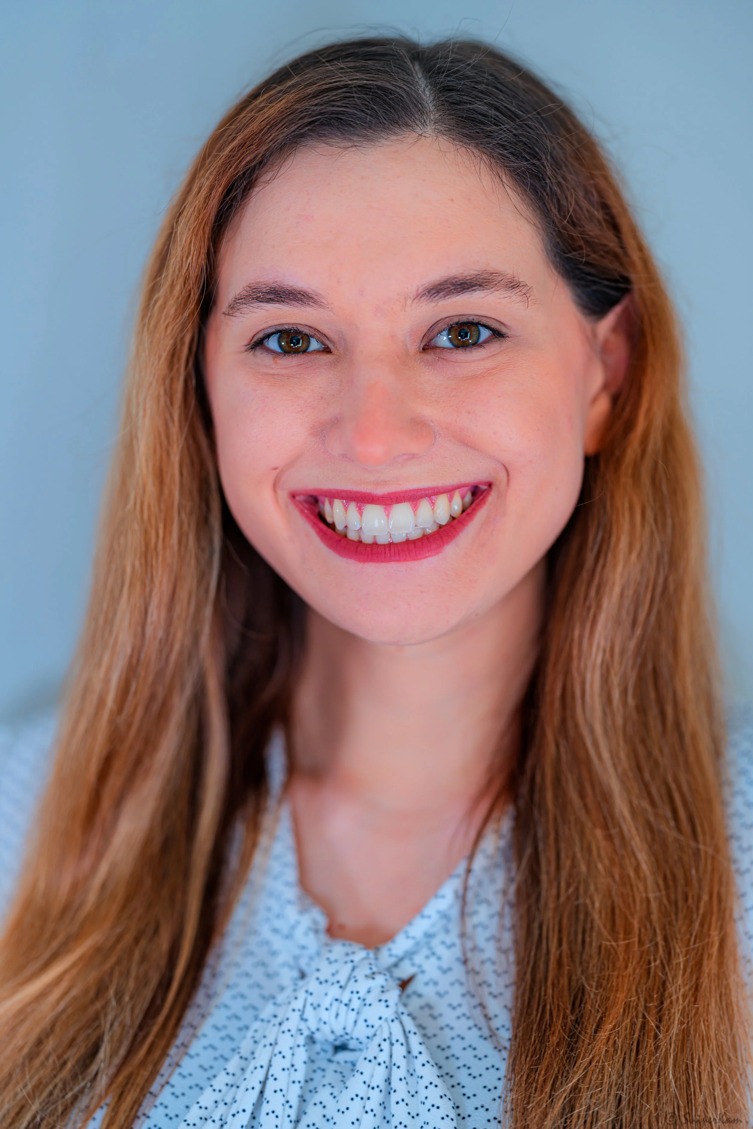 A woman with long, light brown hair, smiling, showing her teeth, against a plain light blue background.