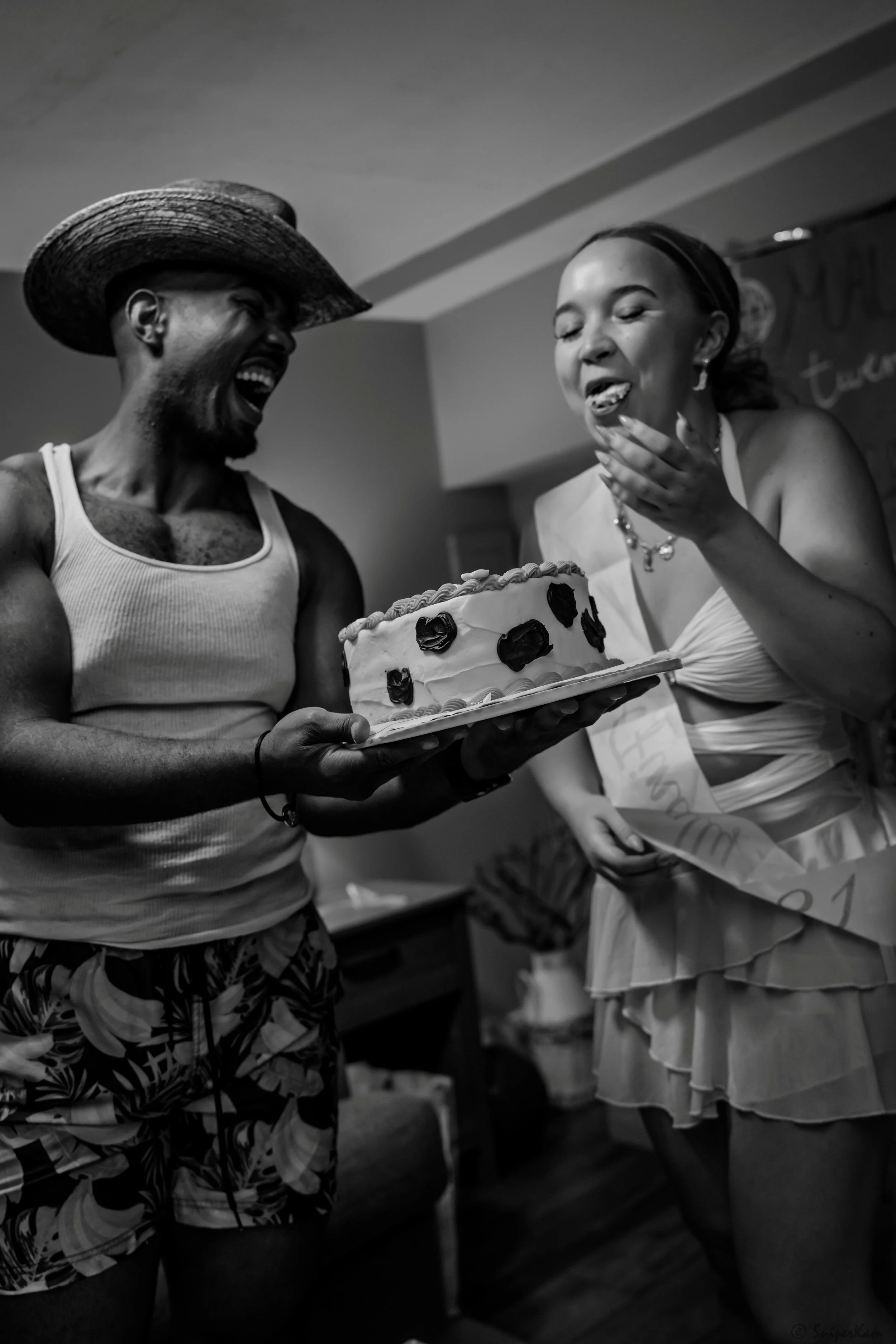 A man in a straw hat and tank top presenting a cake to a woman in a white dress who is wearing a sash, both celebrating with happy expressions.