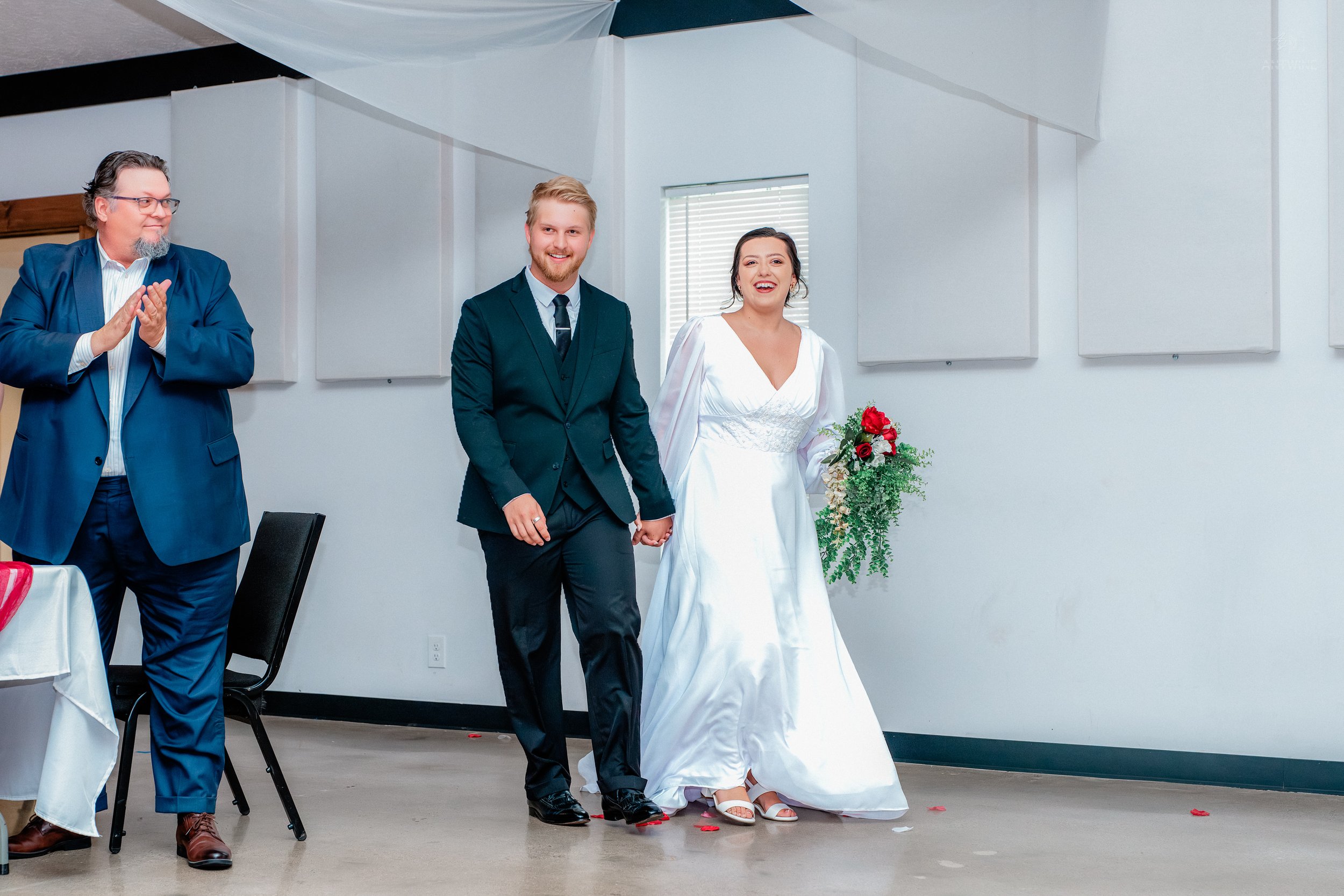 A newly married couple holding hands at their wedding ceremony, standing next to an officiant who is clapping, in a brightly lit room with white walls and windows, with rose petals on the floor.