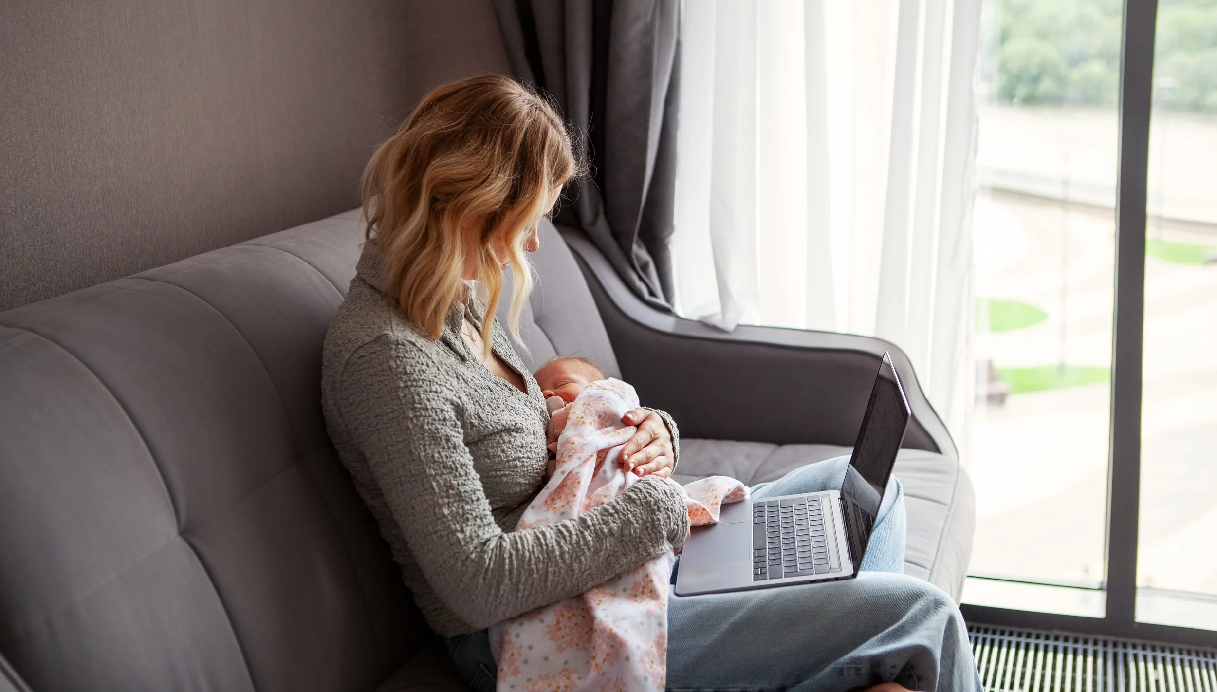A woman is sitting on a gray sofa, breastfeeding a baby wrapped in a pink blanket, while using a laptop.