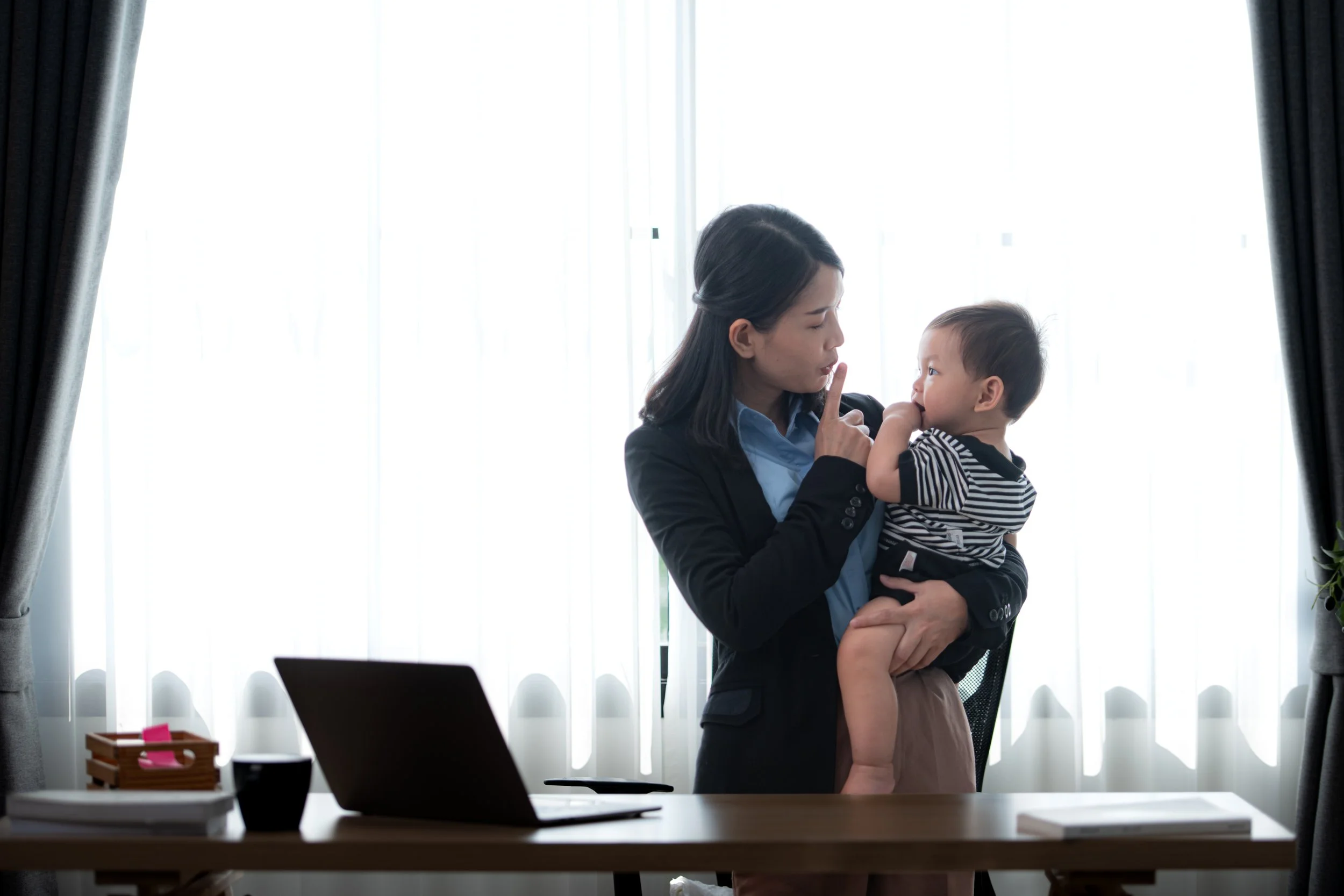 A woman in a business suit holding a young child in an office, making a 'shush' gesture with her finger, standing in front of a desk with a laptop, coffee cup, and office supplies, with natural light coming through curtains.