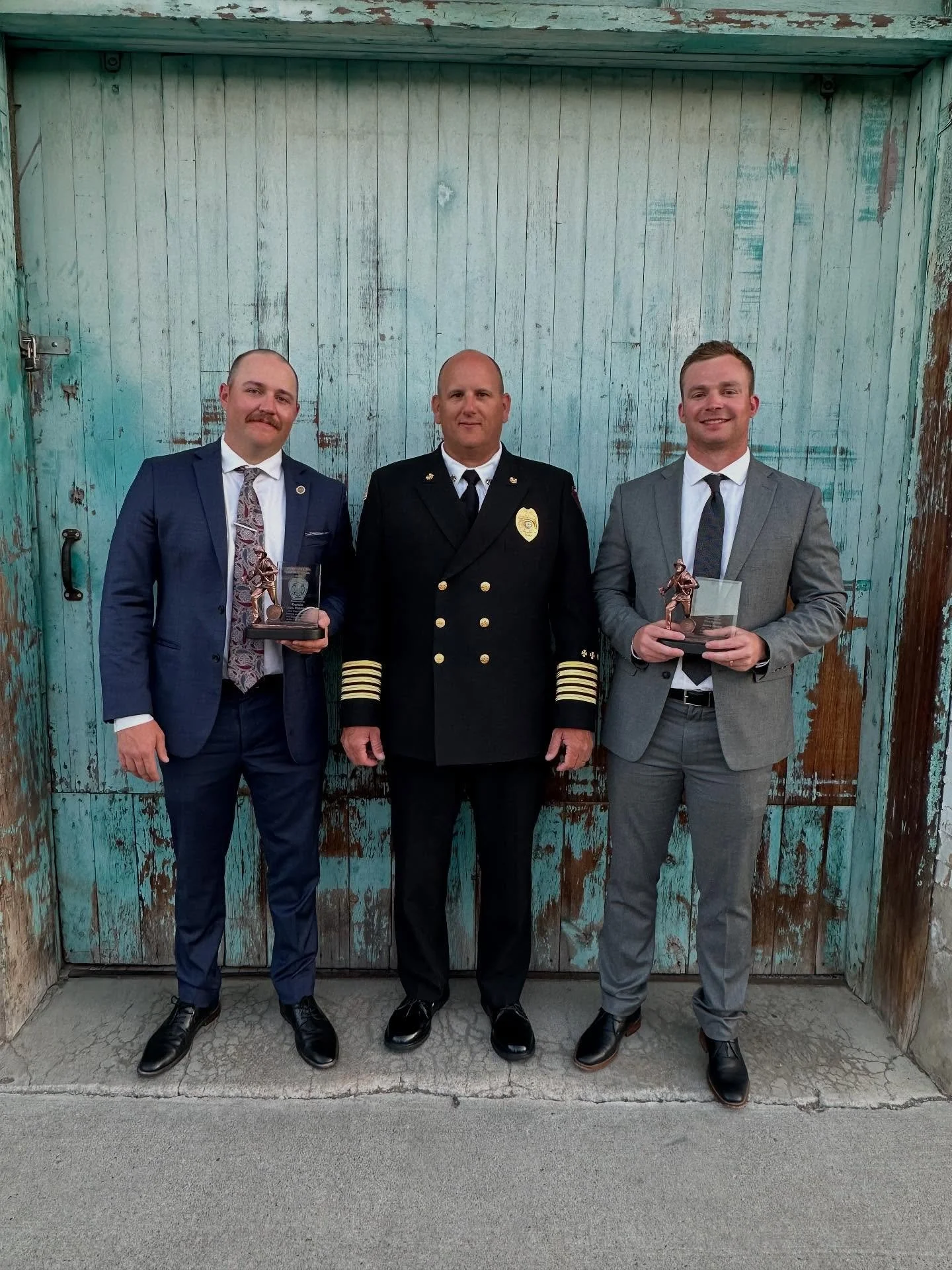 Three men in formal attire standing in front of a weathered, teal-colored wooden door. The man in the middle is in a police uniform, and the two men on either side are holding awards.