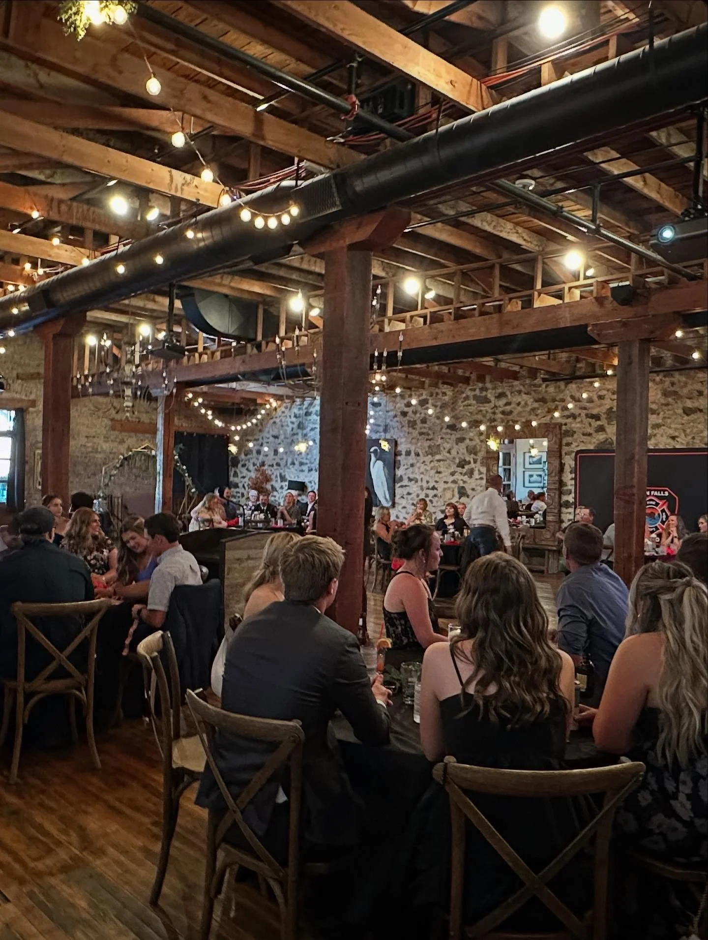 A lively indoor gathering in a rustic-style venue with exposed wooden beams, a stone wall, and string lights. People are seated at tables, engaging in conversation, with a band or speaker on a stage in the background.
