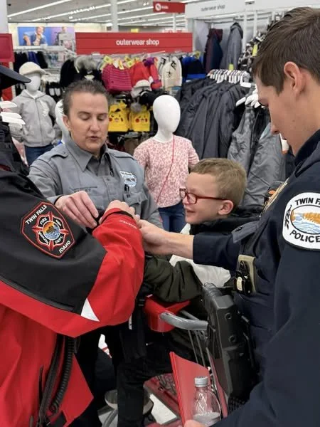 Two police officers, one female and one male, are helping a young boy in a store. The boy is smiling and wearing glasses. The officers are standing next to shopping carts, with the store's clothing section in the background.