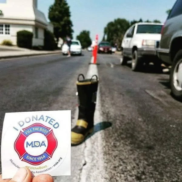 A white hand holding an 'I Donated' sticker with a firefighter logo, a firefighter boot and a traffic cone on the street in the background.