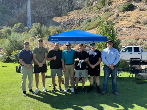 Seven men standing outdoors on grass in front of a blue canopy, with a scenic backdrop of rocks and trees, a pickup truck, and outdoor chairs.