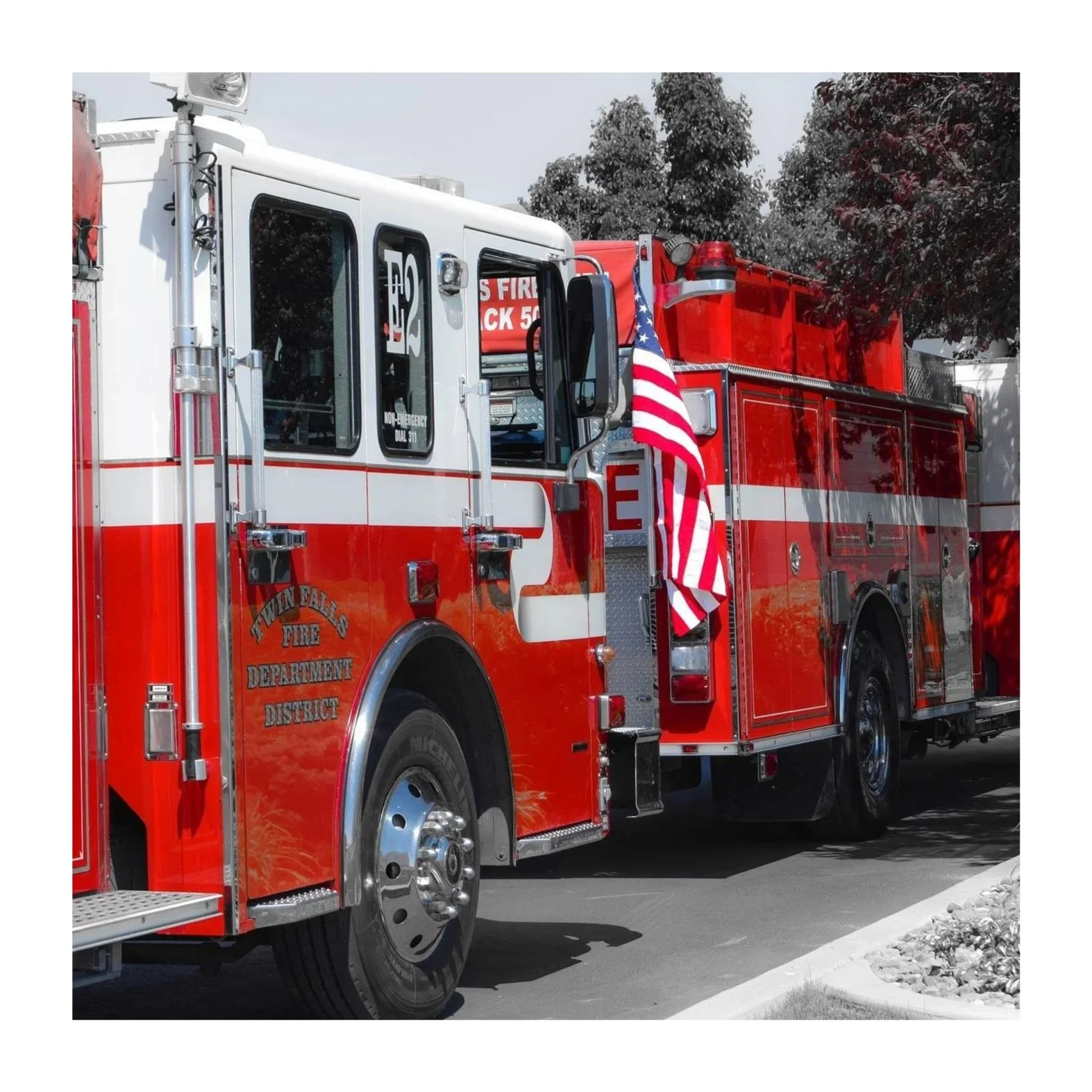A red and white fire truck with an American flag attached, parked on a driveway with trees in the background.