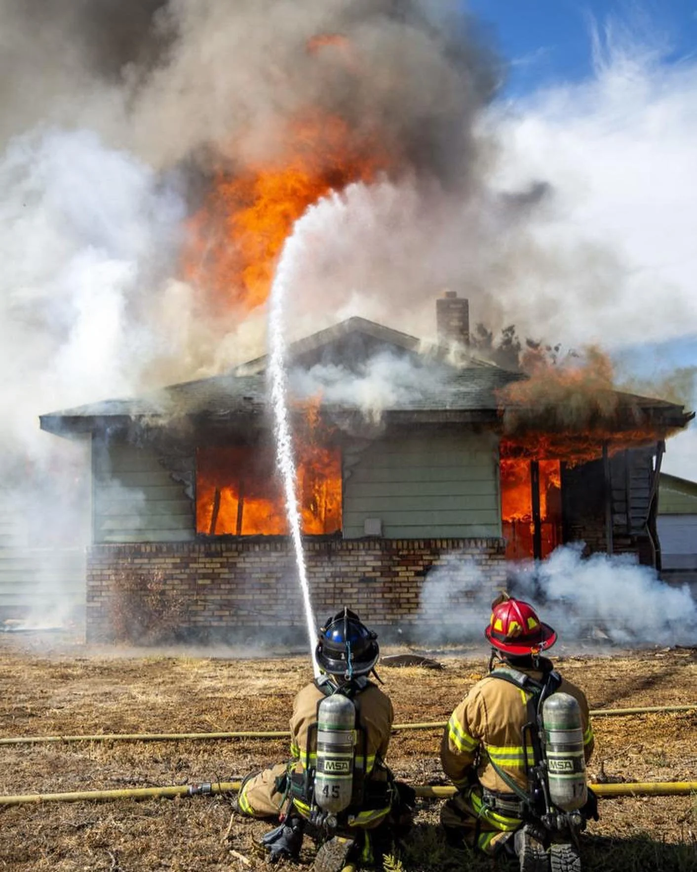 Two firefighters in full gear kneeling in front of a house on fire, spraying water on the flames. The house is engulfed in flames and thick smoke.