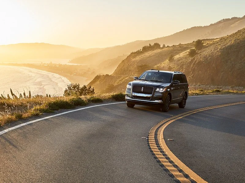 A black SUV driving along a winding coastal road at sunset with hills and the ocean in the background.