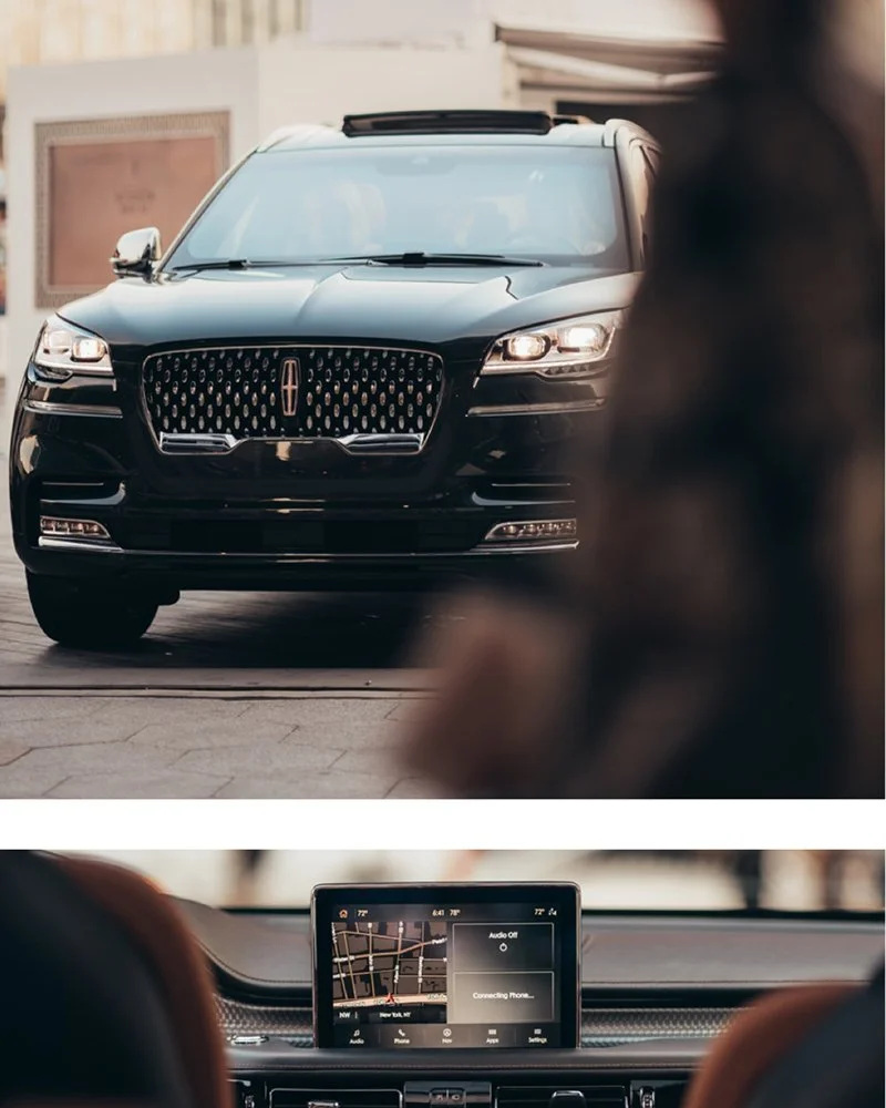 A black luxury SUV with a distinctive grille and headlights parked on a city street, with a person's head visible in the foreground and a car's dashboard with a navigation screen in the background.