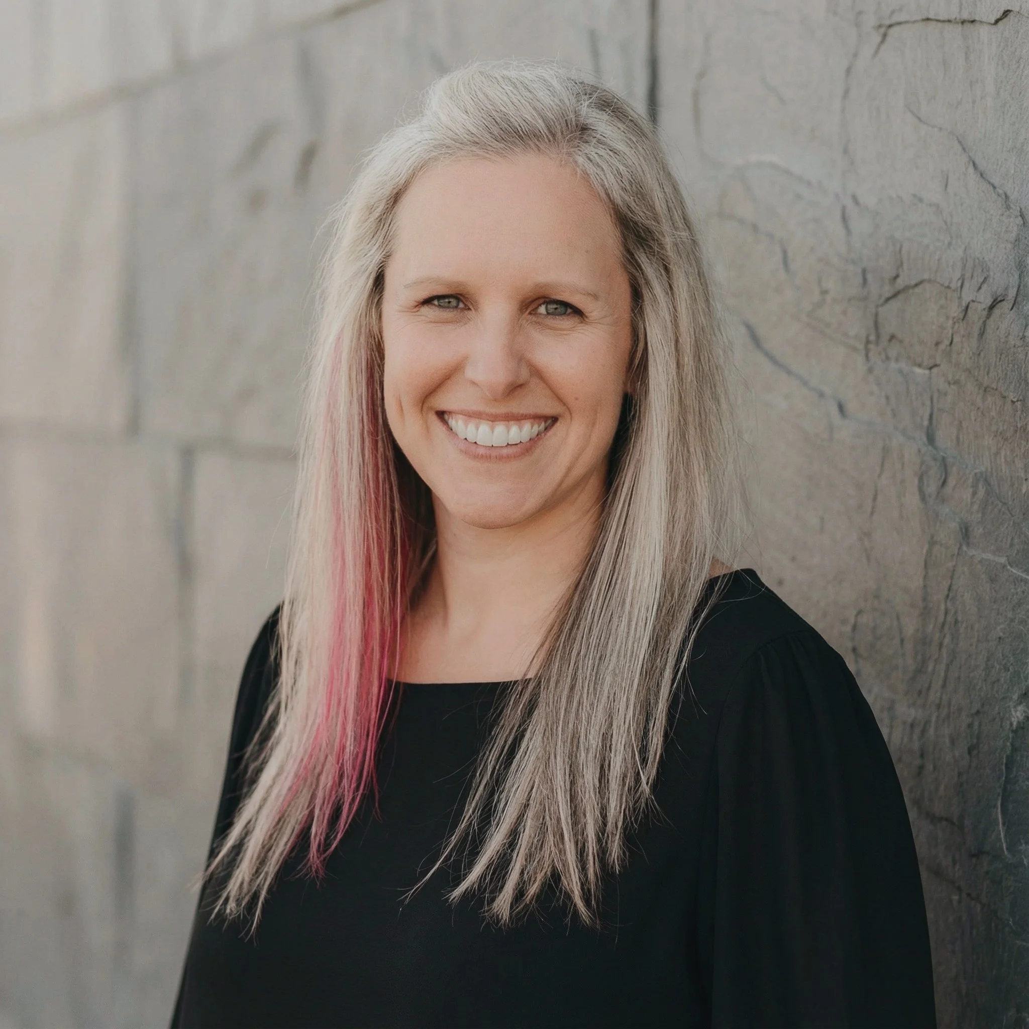 A woman with long, blond hair with pink streaks, smiling, wearing a black top, standing against a gray stone wall.