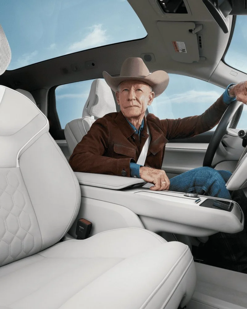 An elderly man wearing a cowboy hat and a brown jacket sitting in the driver's seat of a modern vehicle, with a clear sky visible through the windshield.