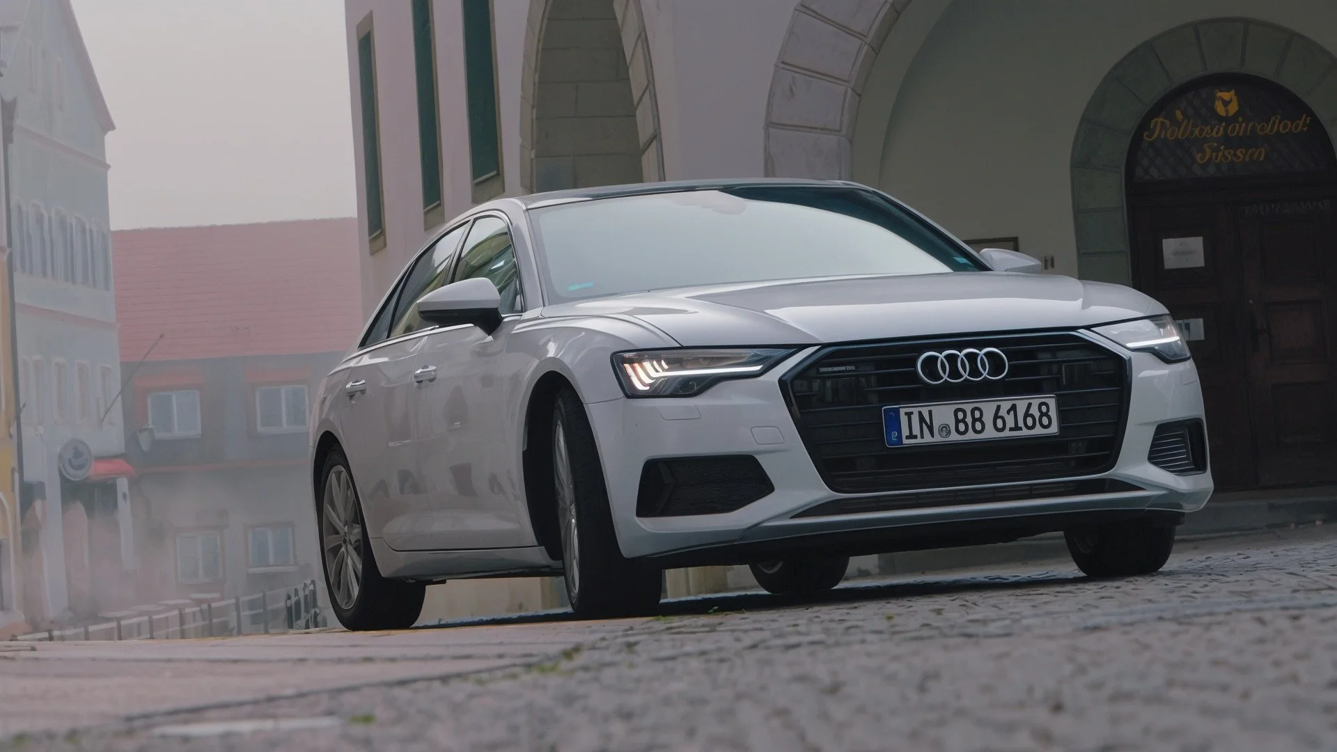 A white Audi sedan parked on a cobblestone street in front of a building with arched entrance and windows, with foggy weather in the background.