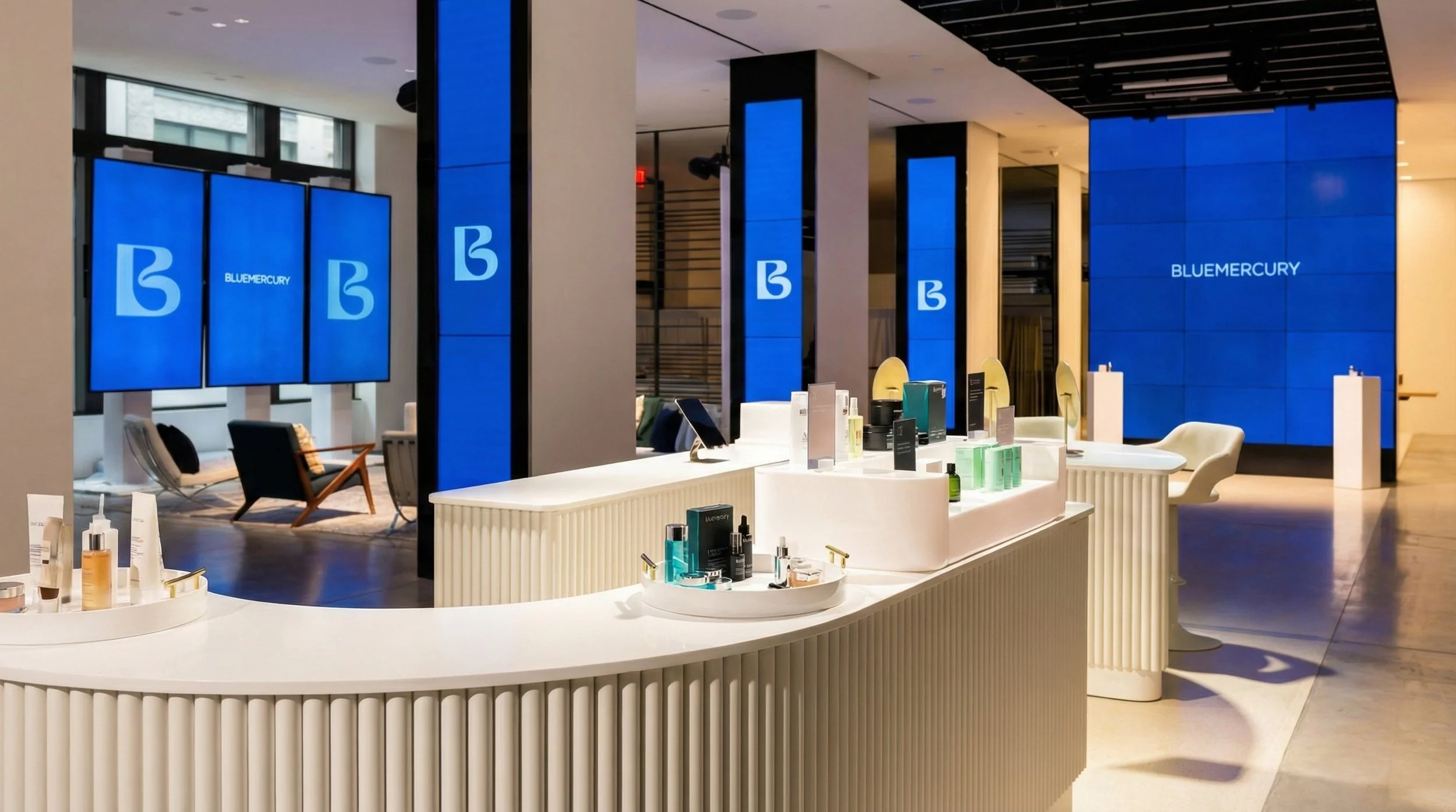 Modern boutique store interior with white display counter featuring skincare and beauty products, blue digital screens with 'BLUE MERCURY' logo, seating area with chairs and couch, and large digital wall display reading 'BLUEMERCURY'.