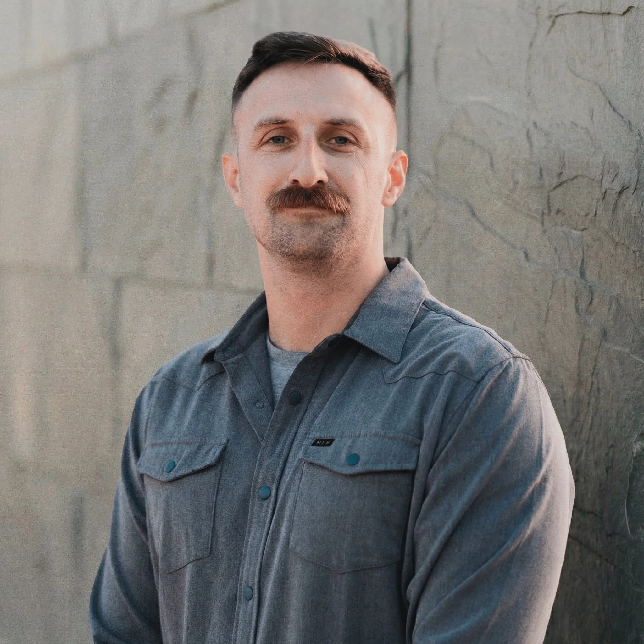 A man with short dark hair, a mustache, and a beard wears a gray denim shirt and leans against a textured stone wall, looking at the camera.