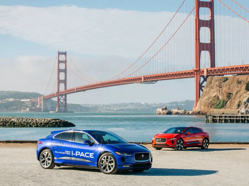 Two electric cars, a blue Jaguar I-PACE and an orange Jaguar model, parked near the San Francisco Bay with the Golden Gate Bridge in the background.