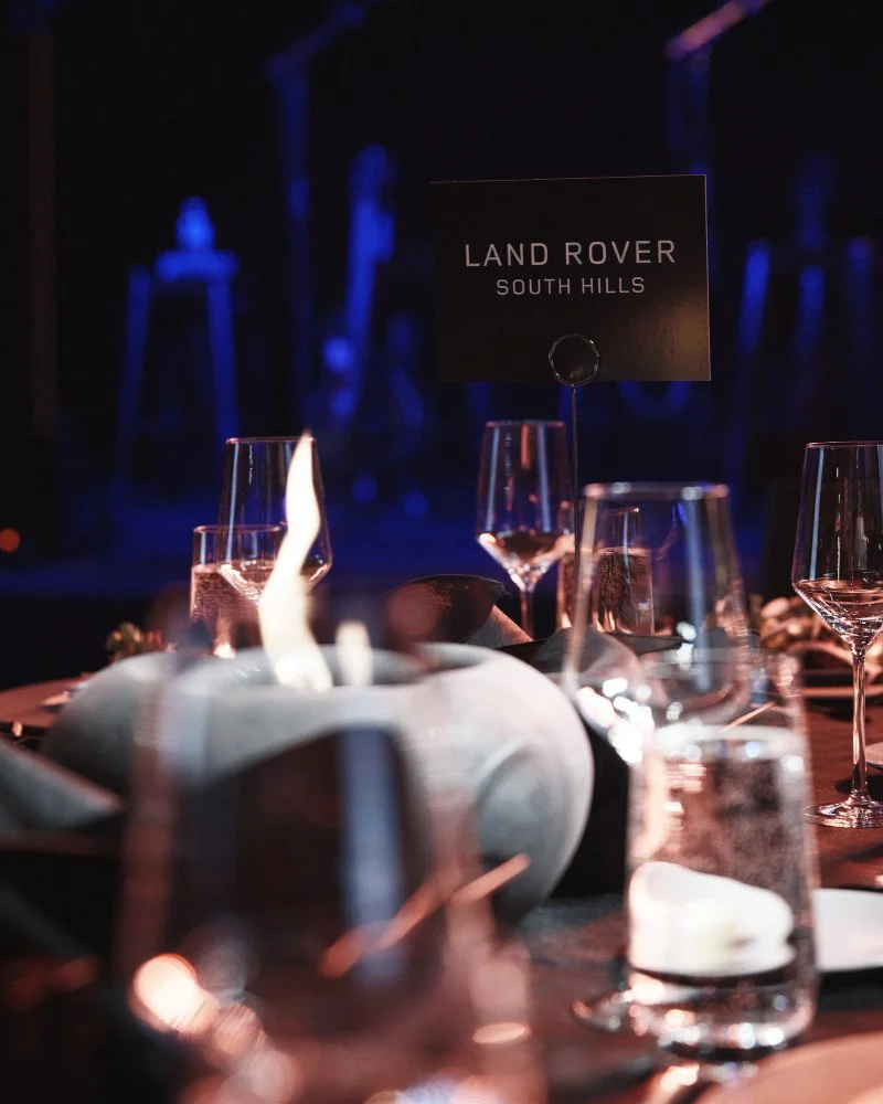 A table at a formal event with wine glasses, a dining candle, and a sign reading 'Land Rover South Hills' in the center.