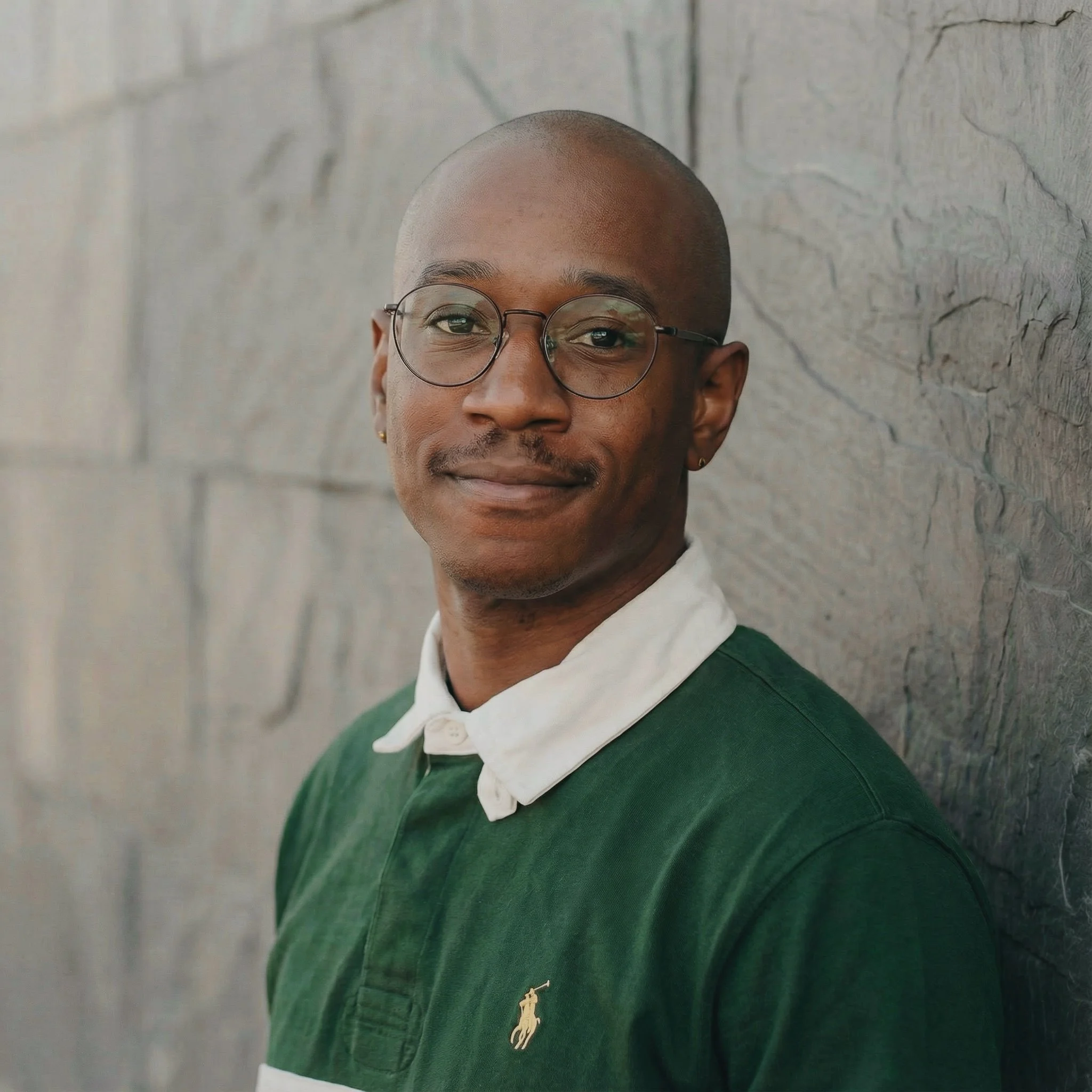 A young man with glasses, a light mustache, and earring, wearing a green Polo shirt with a white collar, leaning against a textured gray wall, looking at the camera with a slight smile.