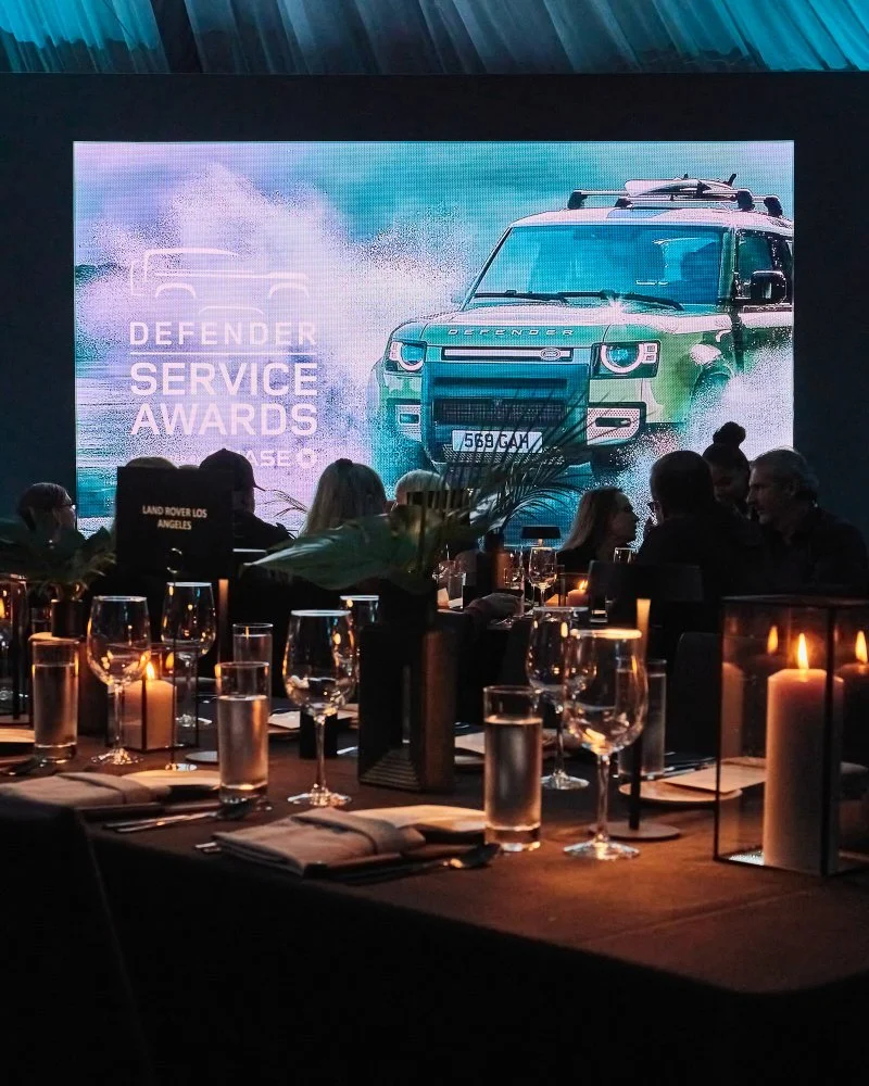 An indoor event with a large screen displaying a Land Rover Defender at a service awards ceremony. The event has a decorated table with candles, glassware, and napkins, with people seated and watching the presentation.