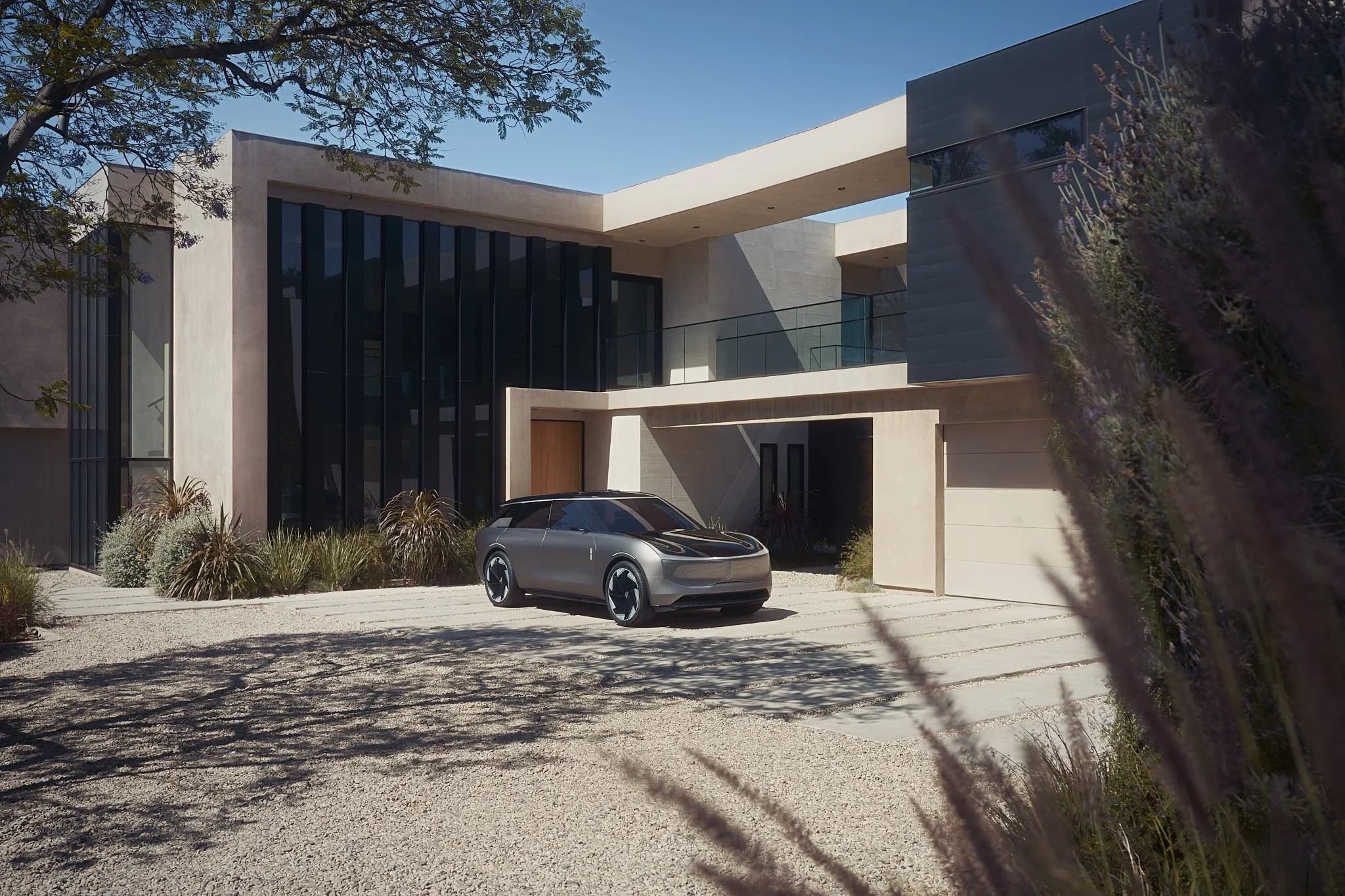 Modern house with large glass windows, beige exterior walls, and a driveway with a futuristic silver car parked in front. Landscaping includes desert plants and trees.