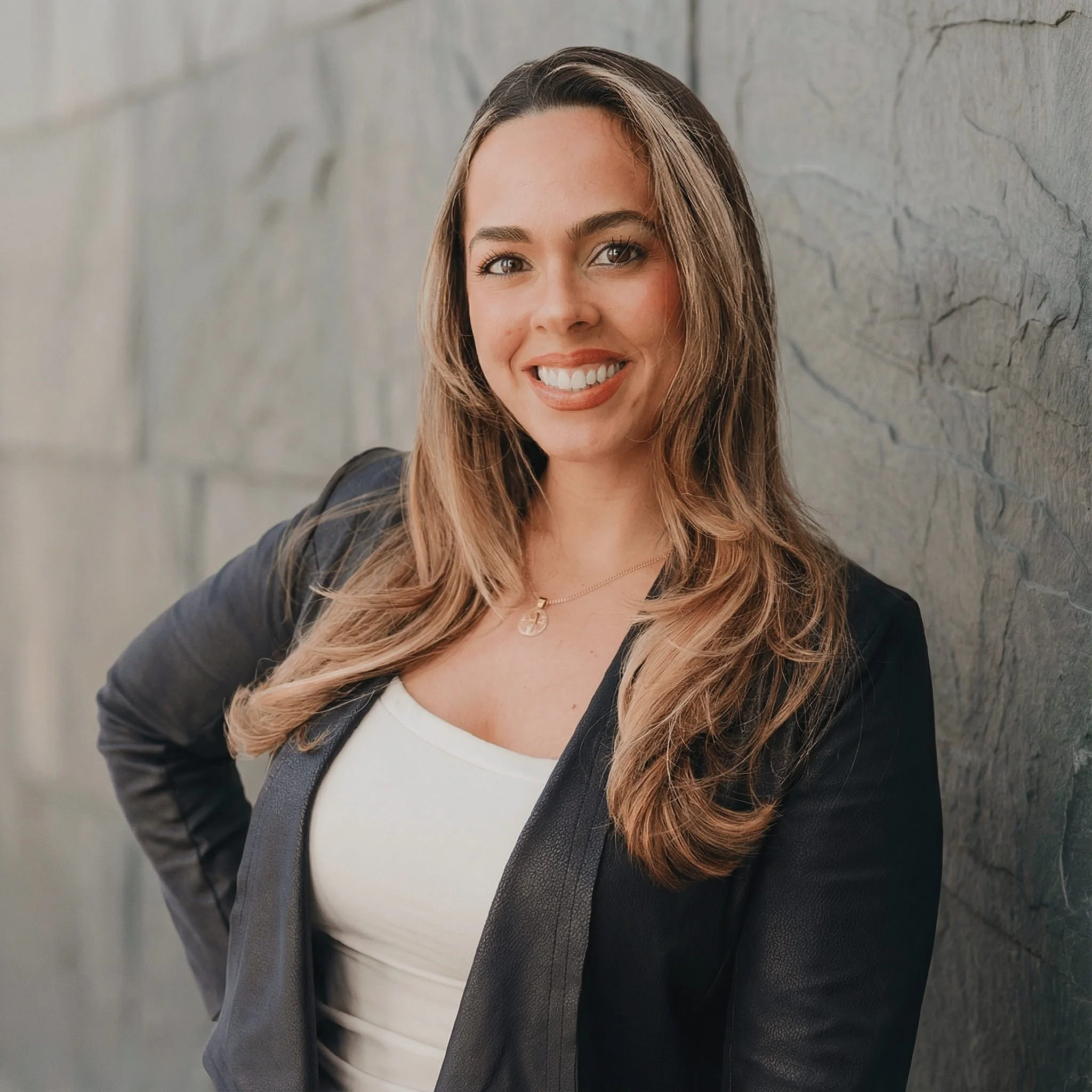 A smiling woman with long, wavy light brown hair, wearing a black blazer over a white top, and a necklace, leaning against a stone wall.