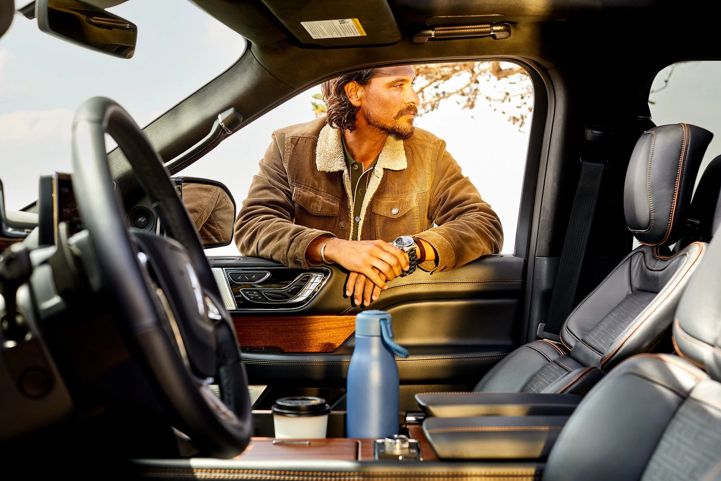 Man leaning on the window of a vehicle, looking outside, with a water bottle and coffee cup on the center console.