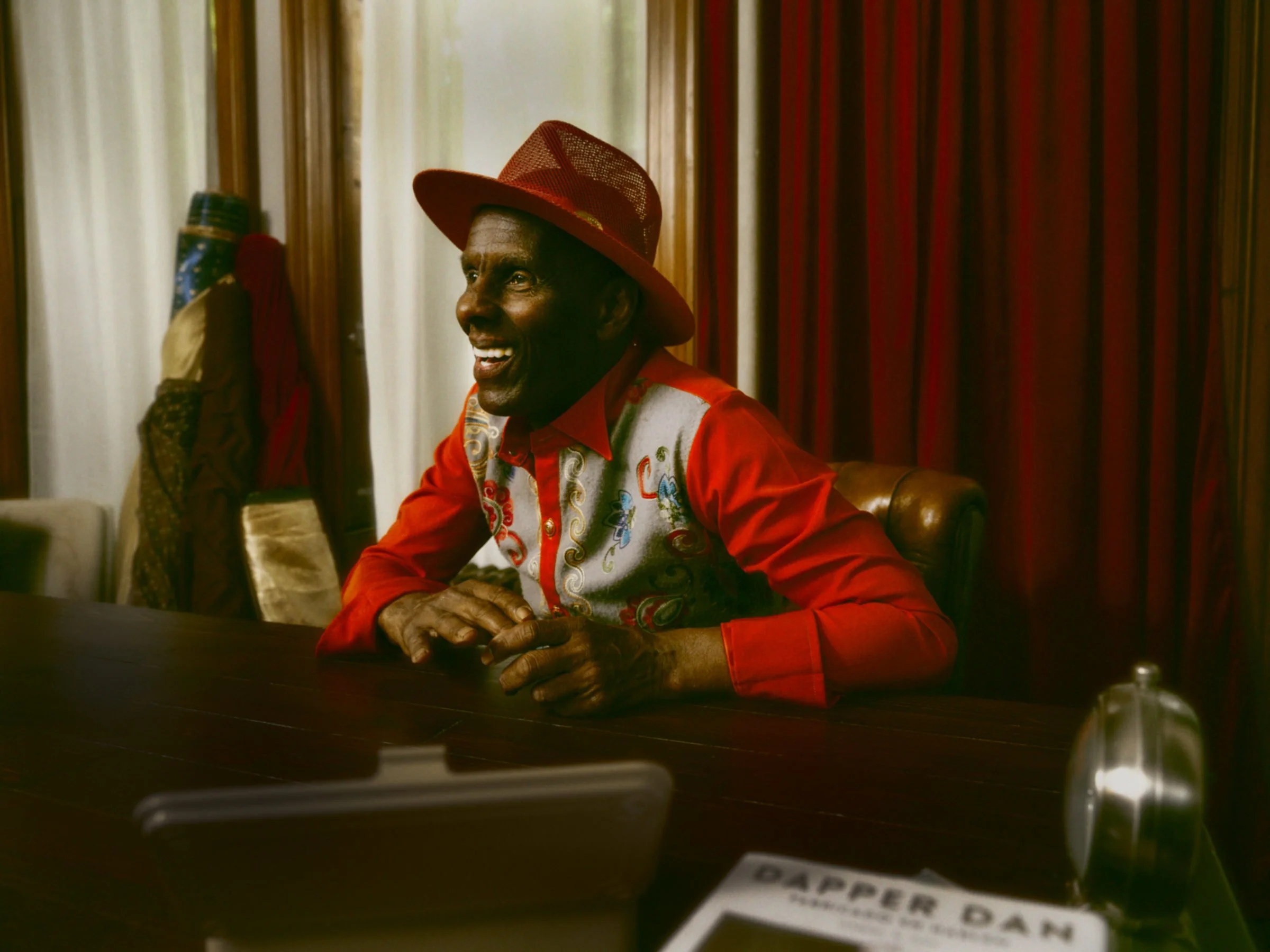 A man wearing a red hat and a colorful embroidered shirt is sitting at a wooden table, smiling and looking to his right. He is in a room with red curtains and wooden paneling.