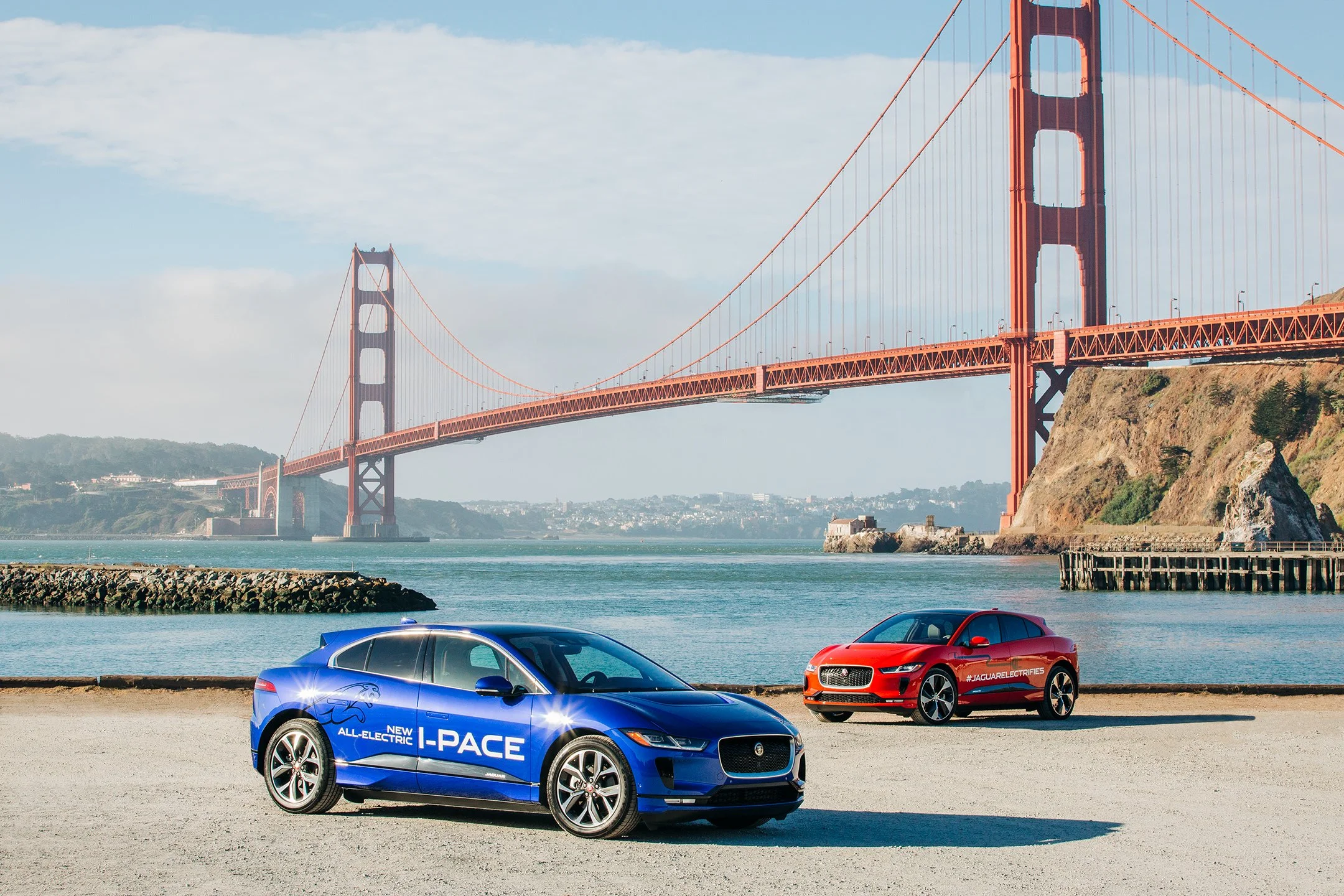 Two Jaguar electric cars parked on a beach near the Golden Gate Bridge in San Francisco, with the city visible in the background.