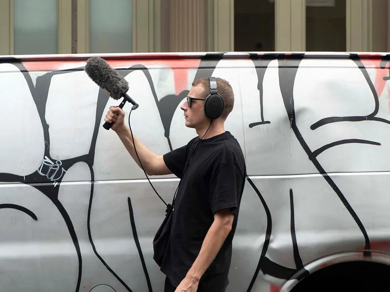 A man wearing sunglasses and headphones is filming graffiti art on the side of a van with a handheld microphone.