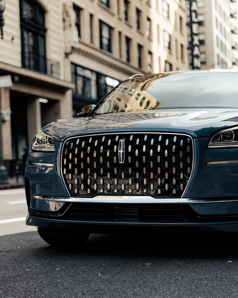 Close-up of the front of a dark luxury car with a distinctive grille, parked on an urban street with tall buildings in the background.