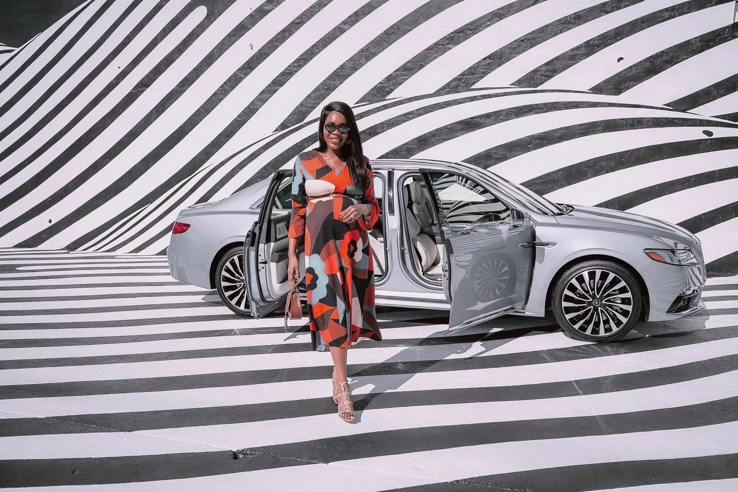 A woman in a colorful floral dress and sunglasses standing in front of a silver luxury car with open doors, set against a black and white striped background.