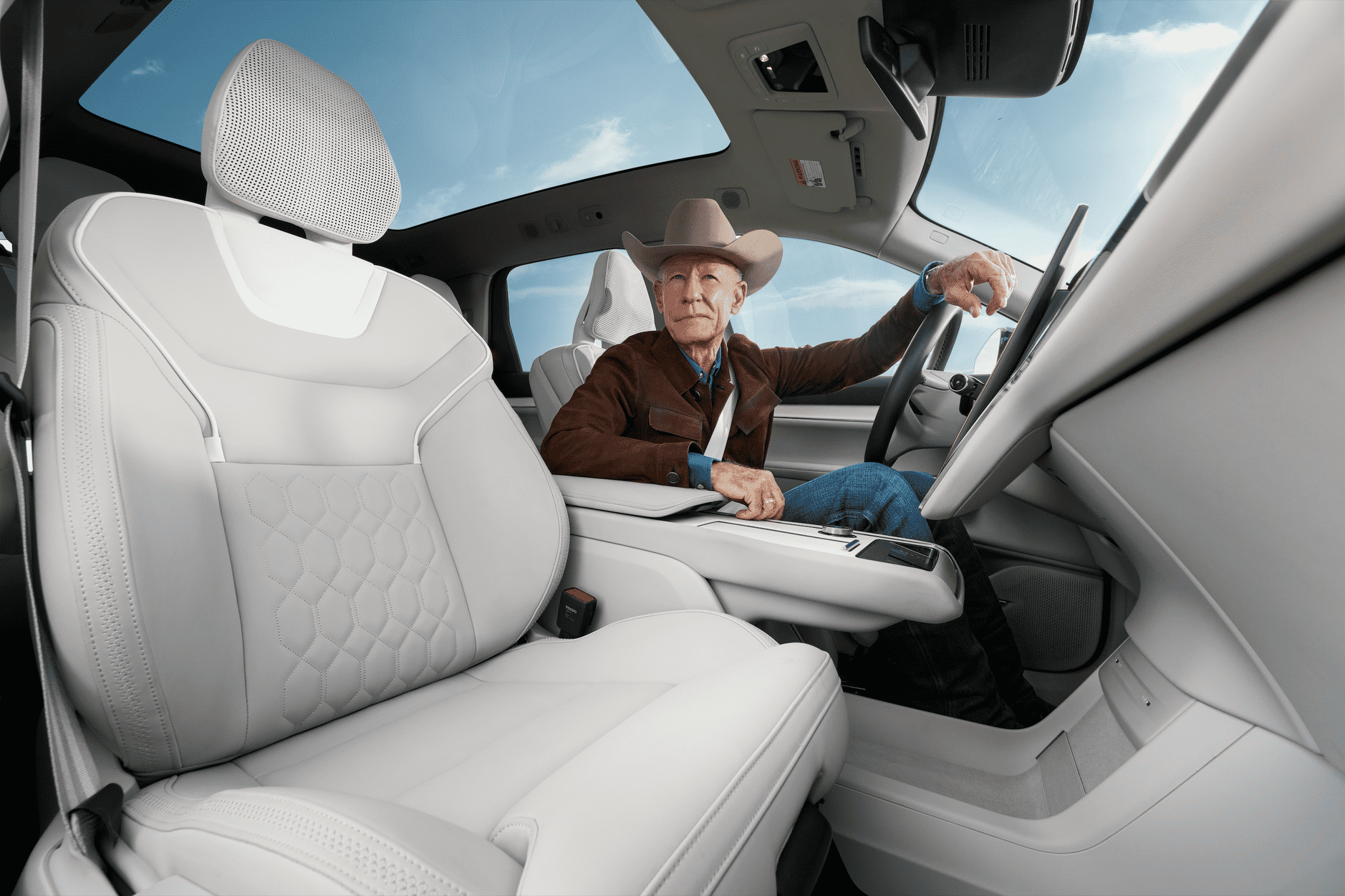 An elderly man wearing a cowboy hat and brown jacket seated in the driver's seat of a modern electric vehicle with white leather seats and panoramic roof, looking at the camera.
