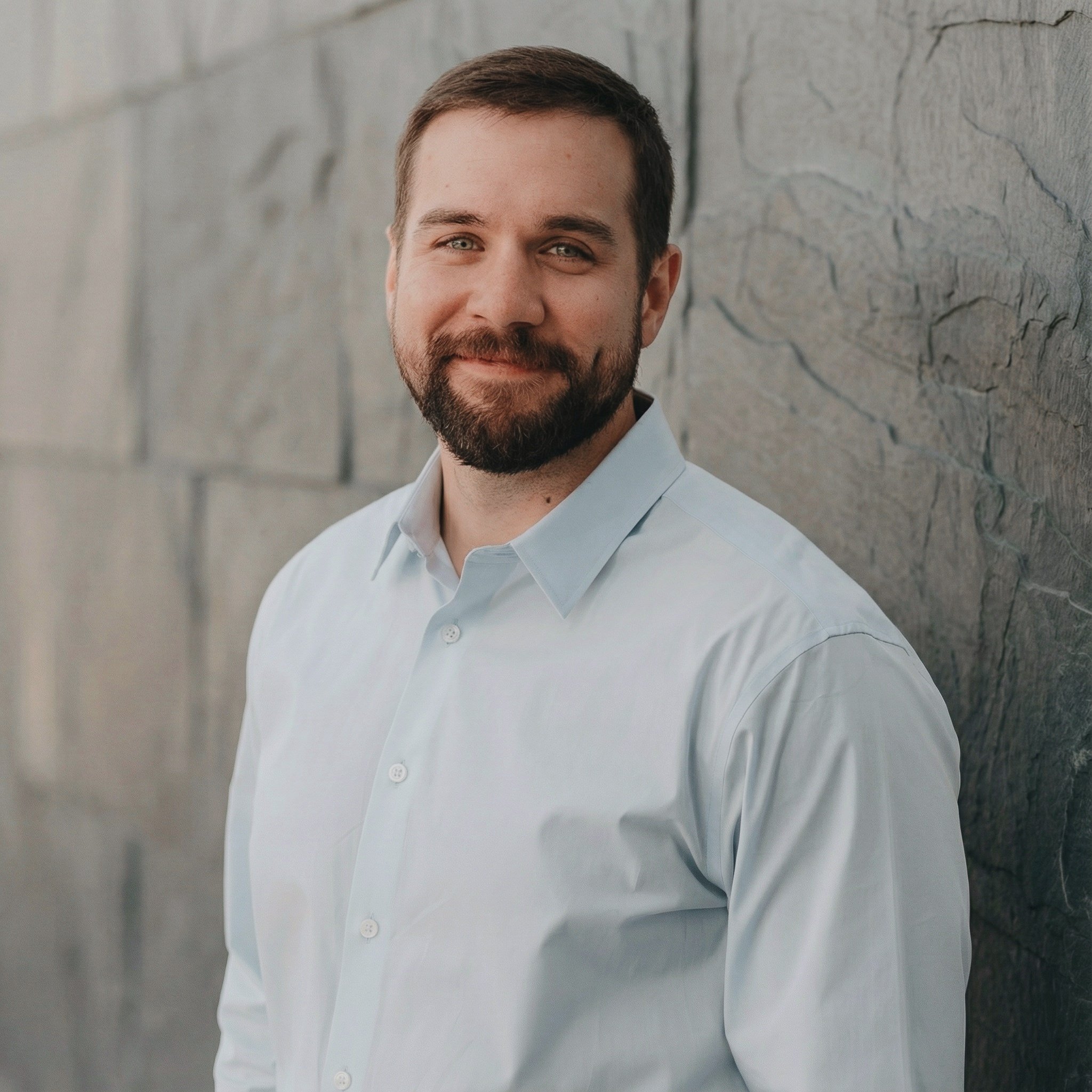 A man with short brown hair, a beard, and blue eyes, wearing a light blue button-up shirt, leaning against a stone wall with a subtle smile.