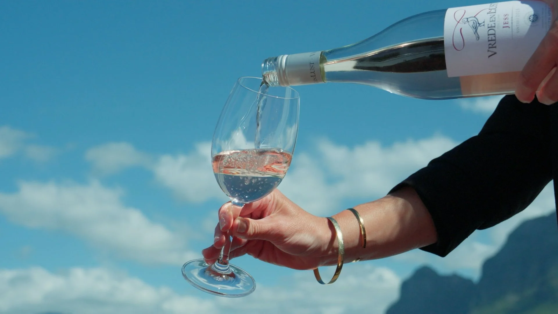A person pouring rosé wine from a bottle into a wine glass outdoors with a blue sky and clouds in the background.