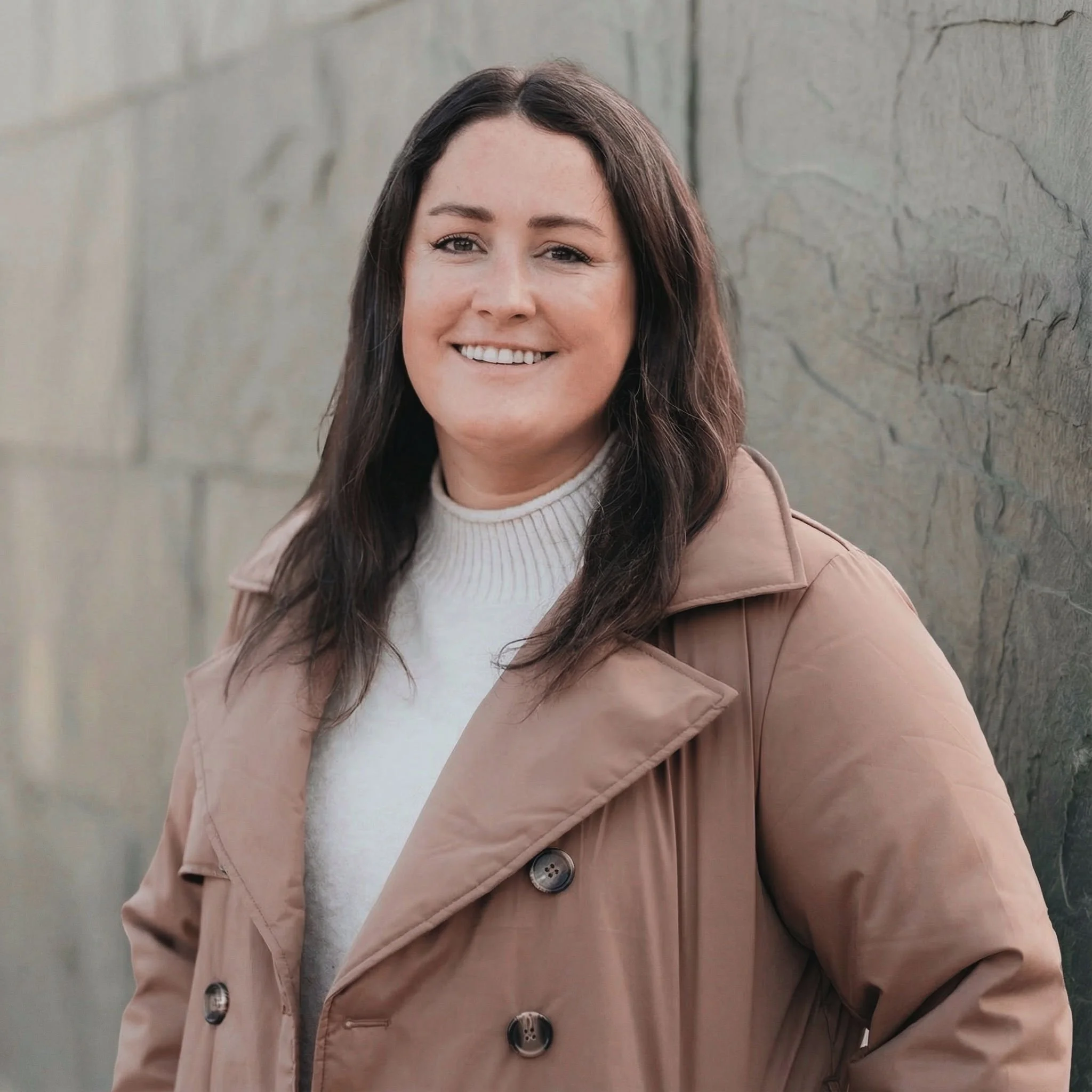 A woman with shoulder-length dark hair smiling, wearing a beige trench coat and a white turtleneck sweater, standing outdoors against a stone wall.