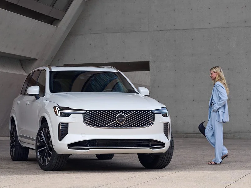 A woman in a light blue suit walking past a white Volvo SUV parked outdoors against a concrete building.
