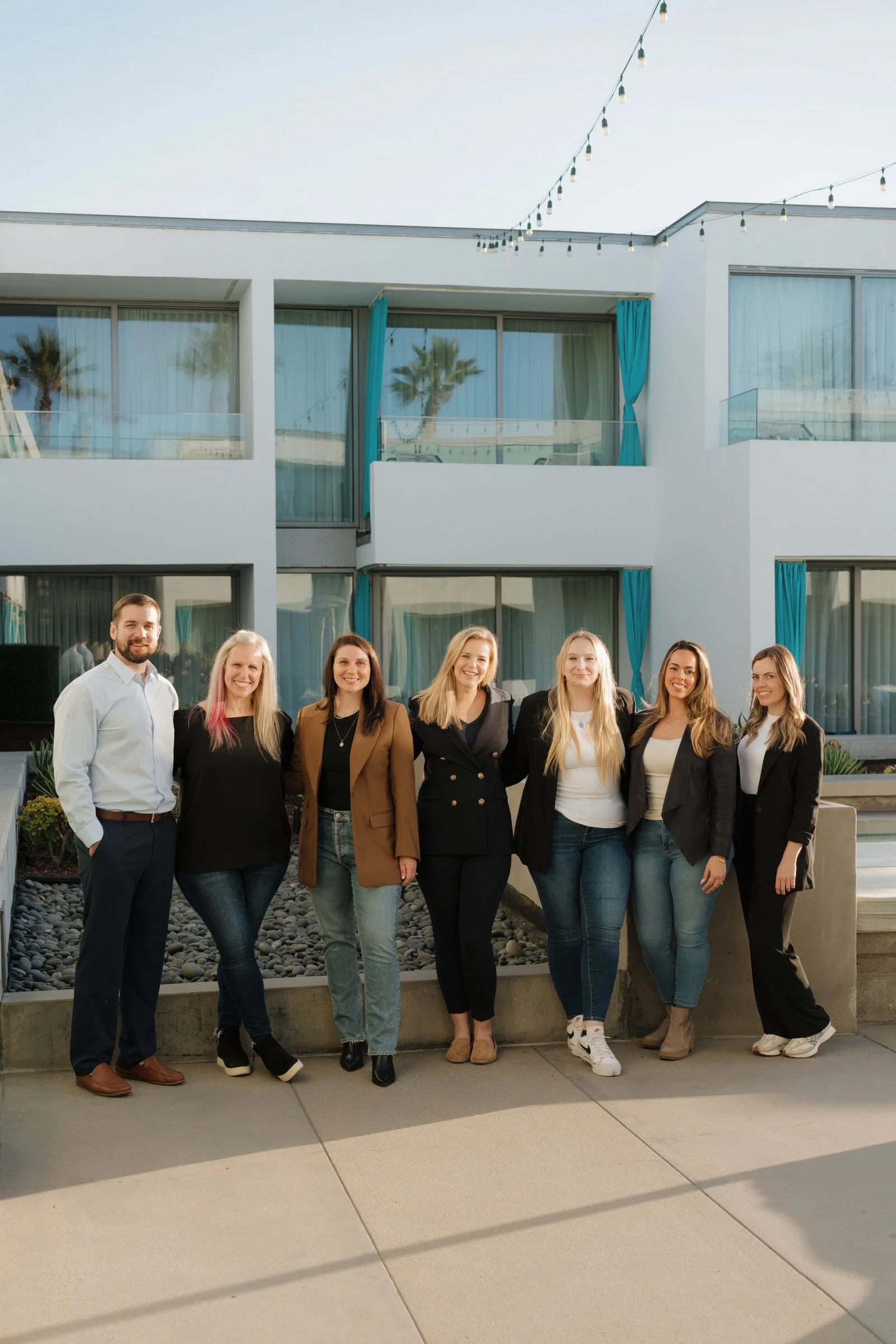 Group of seven people, women and one man, standing outside in front of a modern building with large windows and blue curtains, under string lights, smiling at the camera.