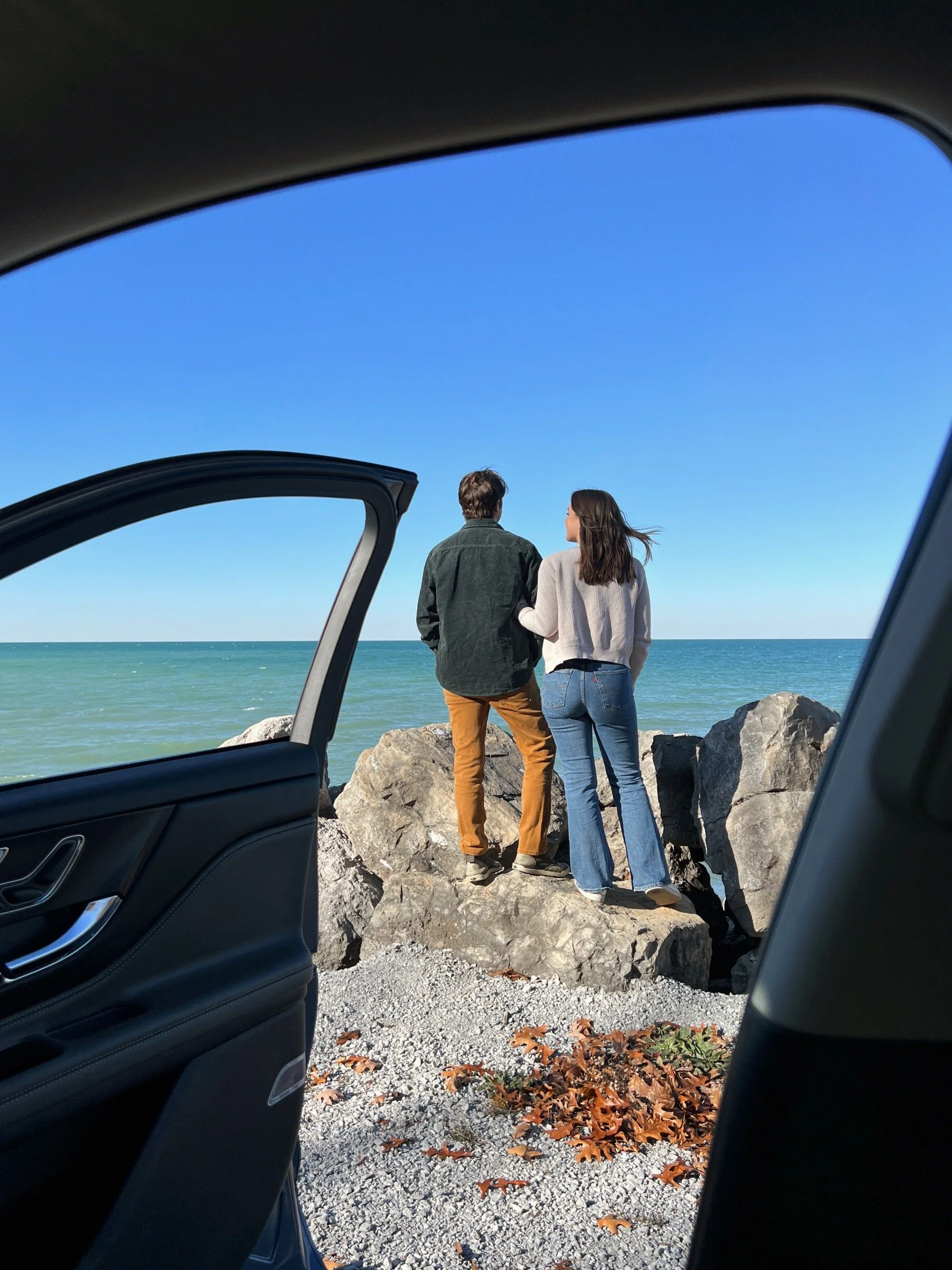 Two people standing on rocks at the beach, viewed from inside a car with the door open, looking out at the ocean under a clear blue sky.