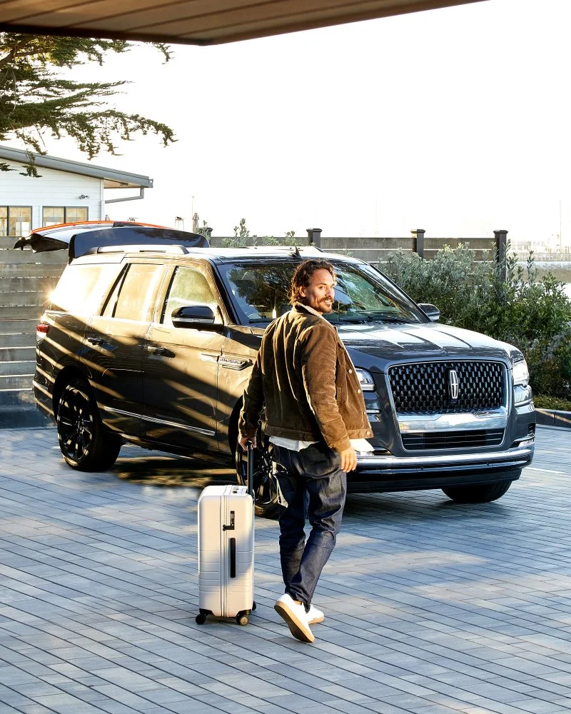 A man with a beard and casual clothing walking with a rolling suitcase in front of a black Lincoln Navigator SUV, outdoors on a wooden deck near a body of water, during sunset.